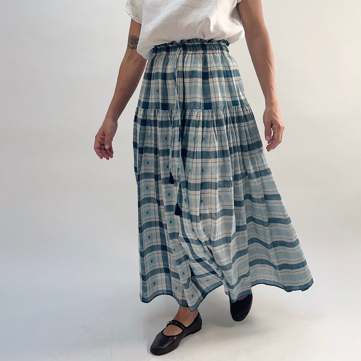 DVAA Denise Skirt in Blue Plaid, tiered cotton maxi skirt in motion with tassel tie swinging, styled with a white tee and black Mary Jane flats — front view mid-stride
