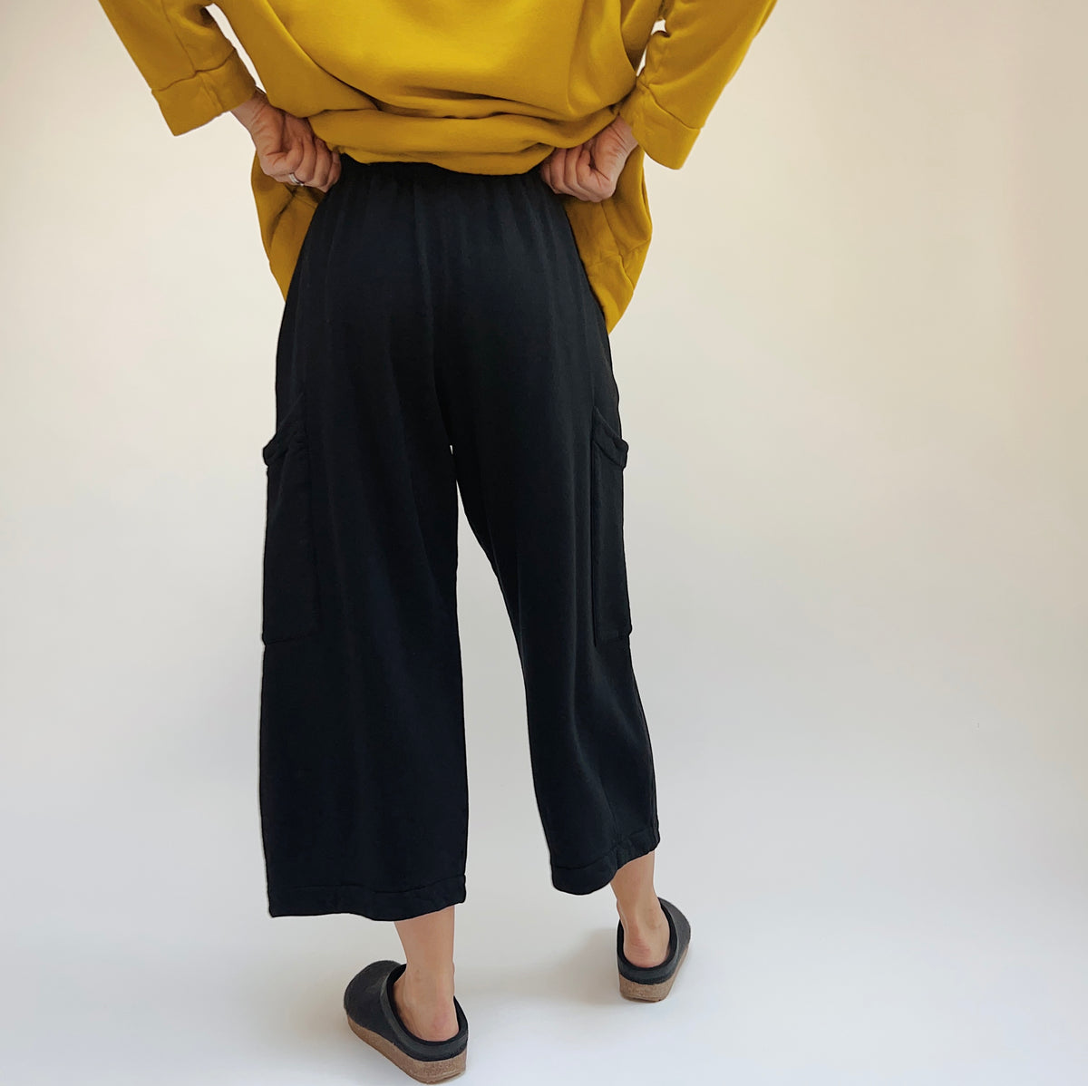 Back lower-body view of a woman wearing the Bryn Walker Pasha Fleece Pant in Black, showing the elastic waistband, back of the thigh patch pockets, wide straight leg silhouette, and folded ankle hem in black organic cotton and bamboo fleece, styled with a mustard yellow sweatshirt and dark slide clogs against a white background
