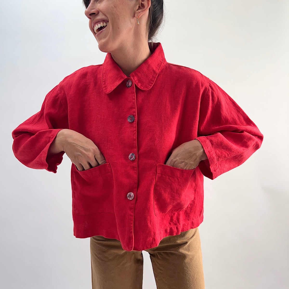 Model smiling and wearing the Bryn Walker Heavy Linen Audrey Jacket in Hawthorn, front view with hands in patch pockets, paired with tan trousers
