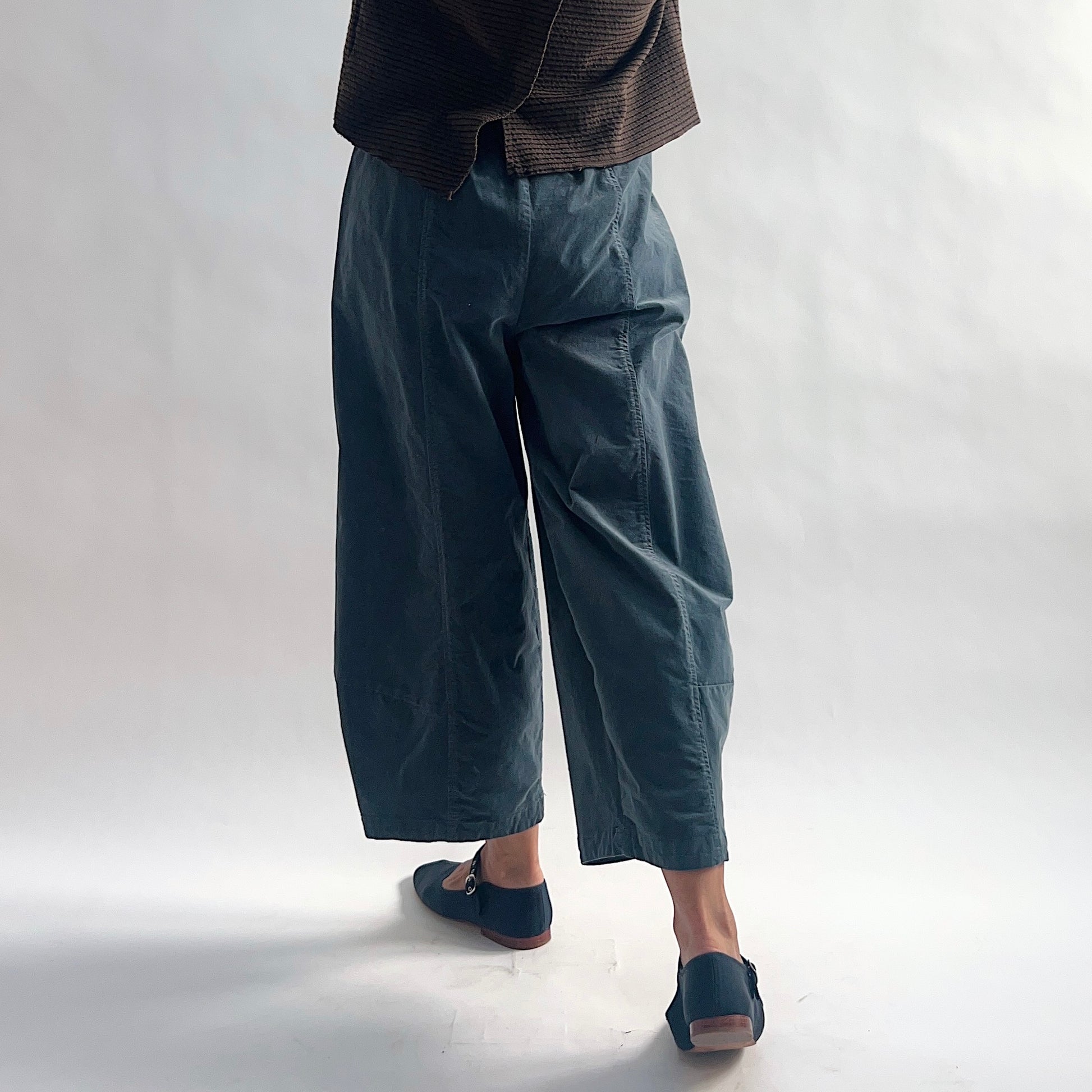 Back view of model wearing iron blue-gray mini cord lantern pants, showing the elastic waist gathering, side seam, wide rounded volume, and tapered ankle, styled with the matching chocolate striped cardigan and blue mary jane flats against a white background.
