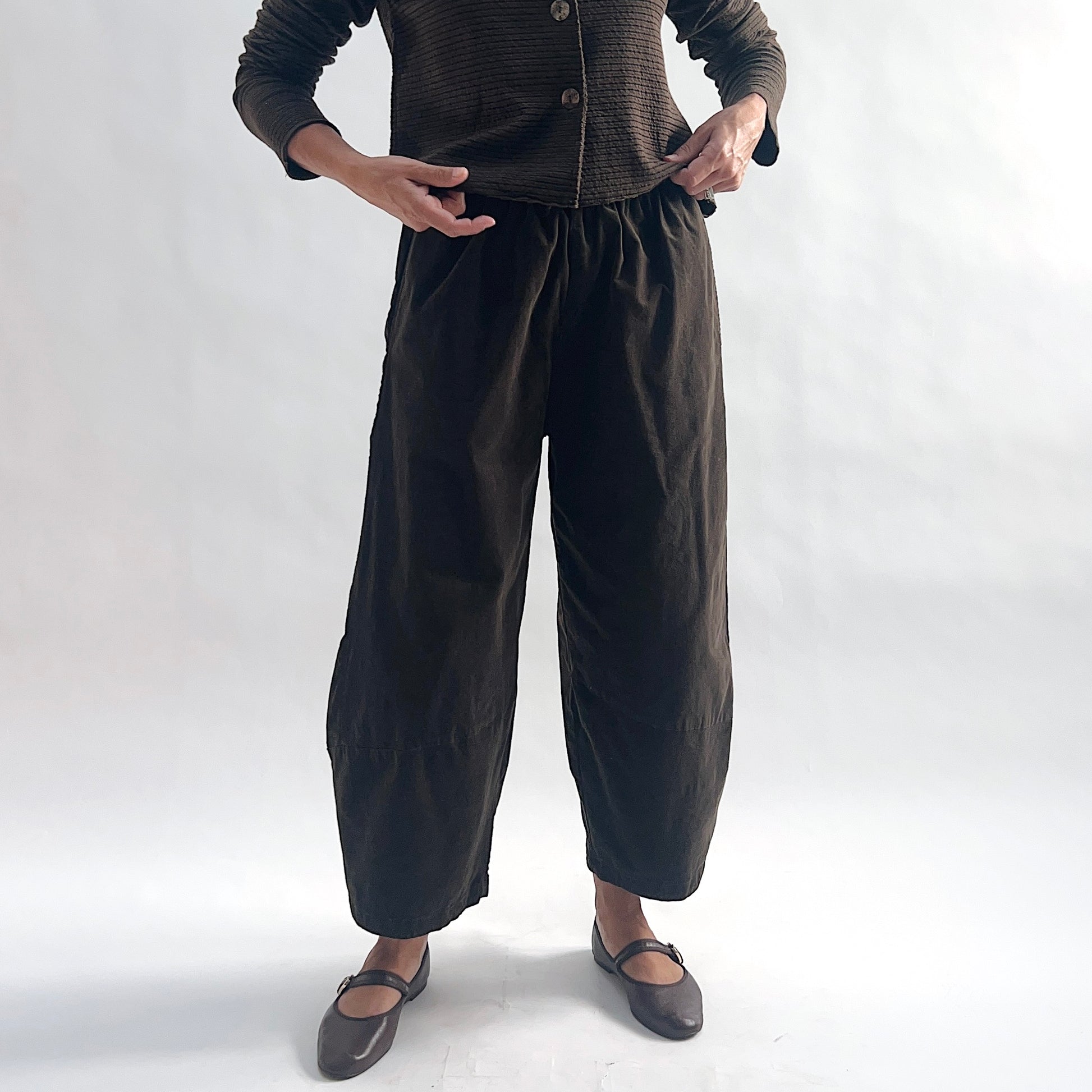 Full front view of model wearing chocolate mini cord lantern pants with elastic gathered waist, hands on hips, showing the wide rounded silhouette and tapered ankle, styled with the matching striped cropped cardigan and dark mary jane flats against a white background.
