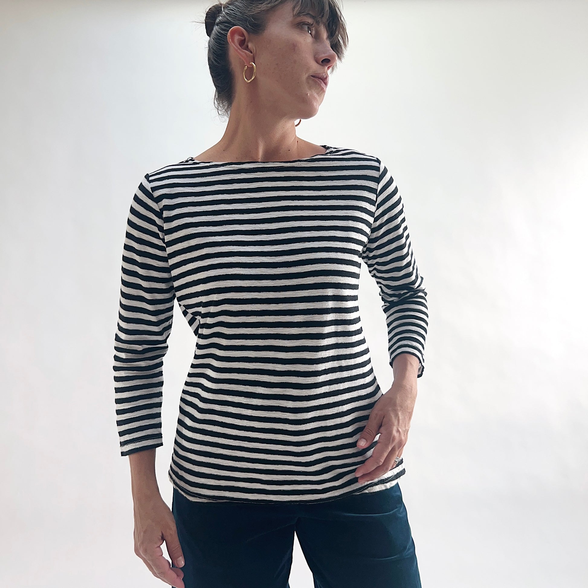 Three-quarter front view of model wearing a navy and white laundered stripe 3/4 sleeve boatneck tee, looking to the side with hand on hip, showing the boatneck neckline and relaxed silhouette, styled with dark blue pants.
