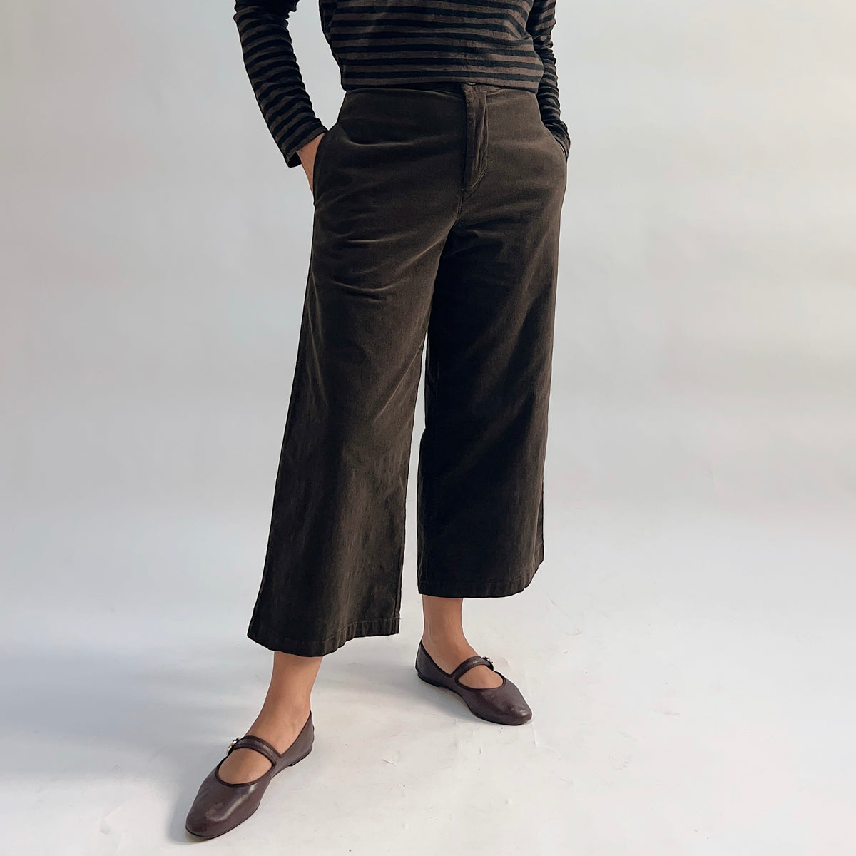 Model wearing the Cut Loose Straight Crop Trouser in Chocolate from a slight angle, showing the wide-leg straight crop silhouette, high waist, and front side pockets in rich dark chocolate cotton-spandex twill, cropped just above the ankle and paired with brown Mary Jane flats and a striped top
