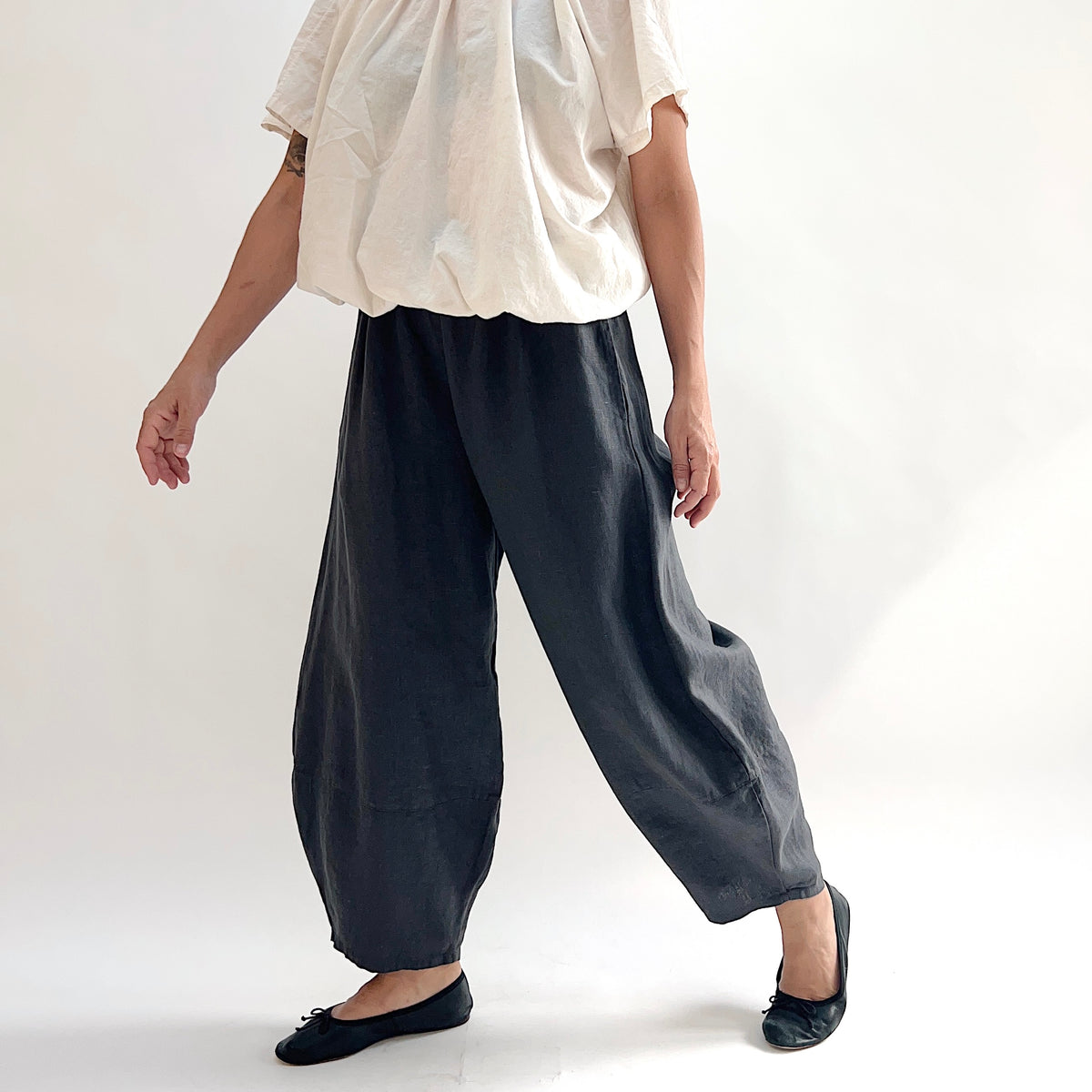 Model in motion wearing the Bryn Walker Heavy Linen Oliver Pant in Moonlight, three-quarter front view showing barrel-leg movement, elastic waistband, and tapered ankle in deep moonlight charcoal heavy linen, styled with cream short-sleeve top and black ballet flats

