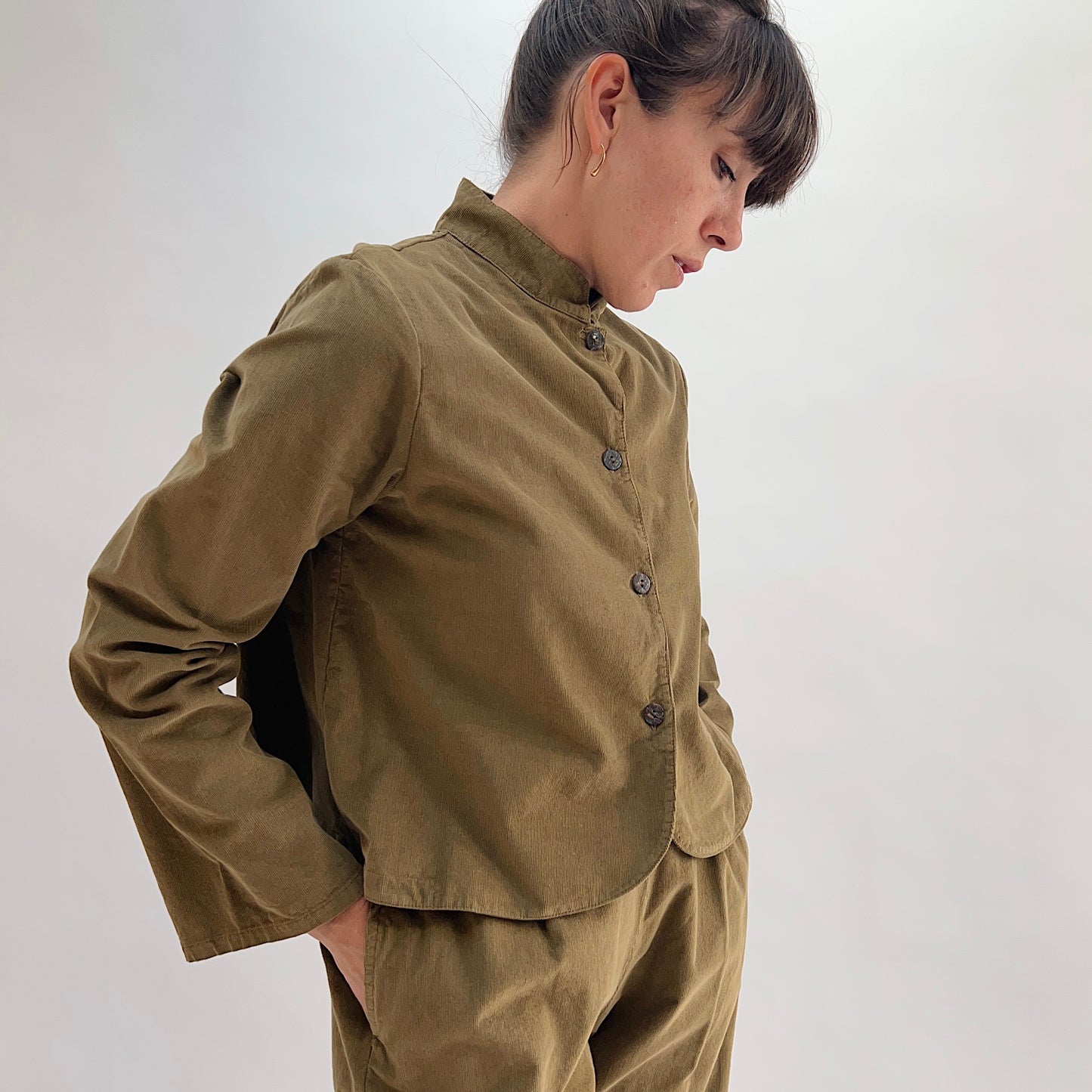 Model looking down wearing the Bryn Walker Corduroy Standing Collar Crop Jacket in Partridge, three-quarter front view highlighting ruffled collar, shell buttons, wide sleeve cuff, and olive pinwheel corduroy, paired with matching Pasha Pants
