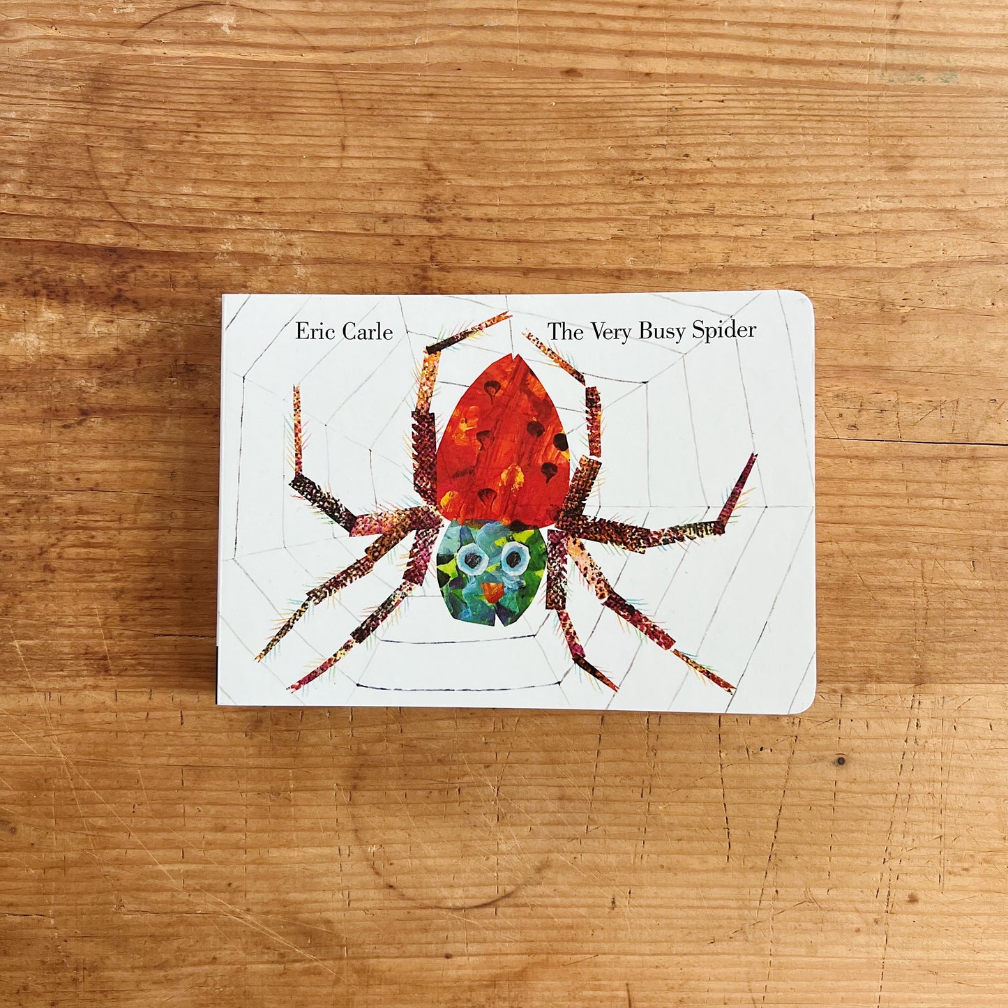 Cover of The Very Busy Spider board book by Eric Carle, featuring a large collage-art spider with a red and orange body centered on a silkweb against a white background, with author and title text in the upper corners
