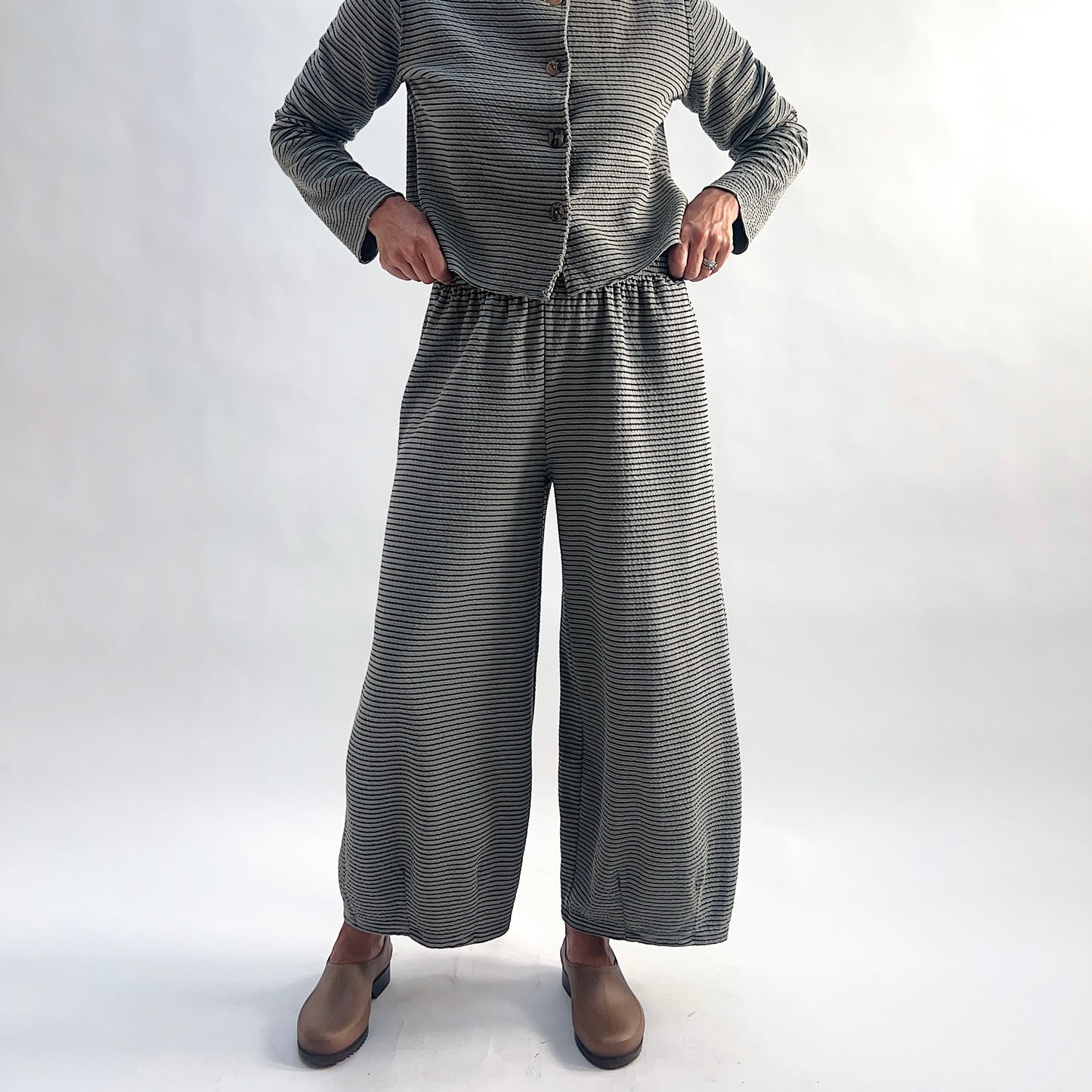 Front view of model wearing squirrel and black fine horizontal stripe wide-leg cropped pants with elastic waist and dart shaping, styled with the matching striped cropped cardigan and taupe slip-on shoes against a white background.
