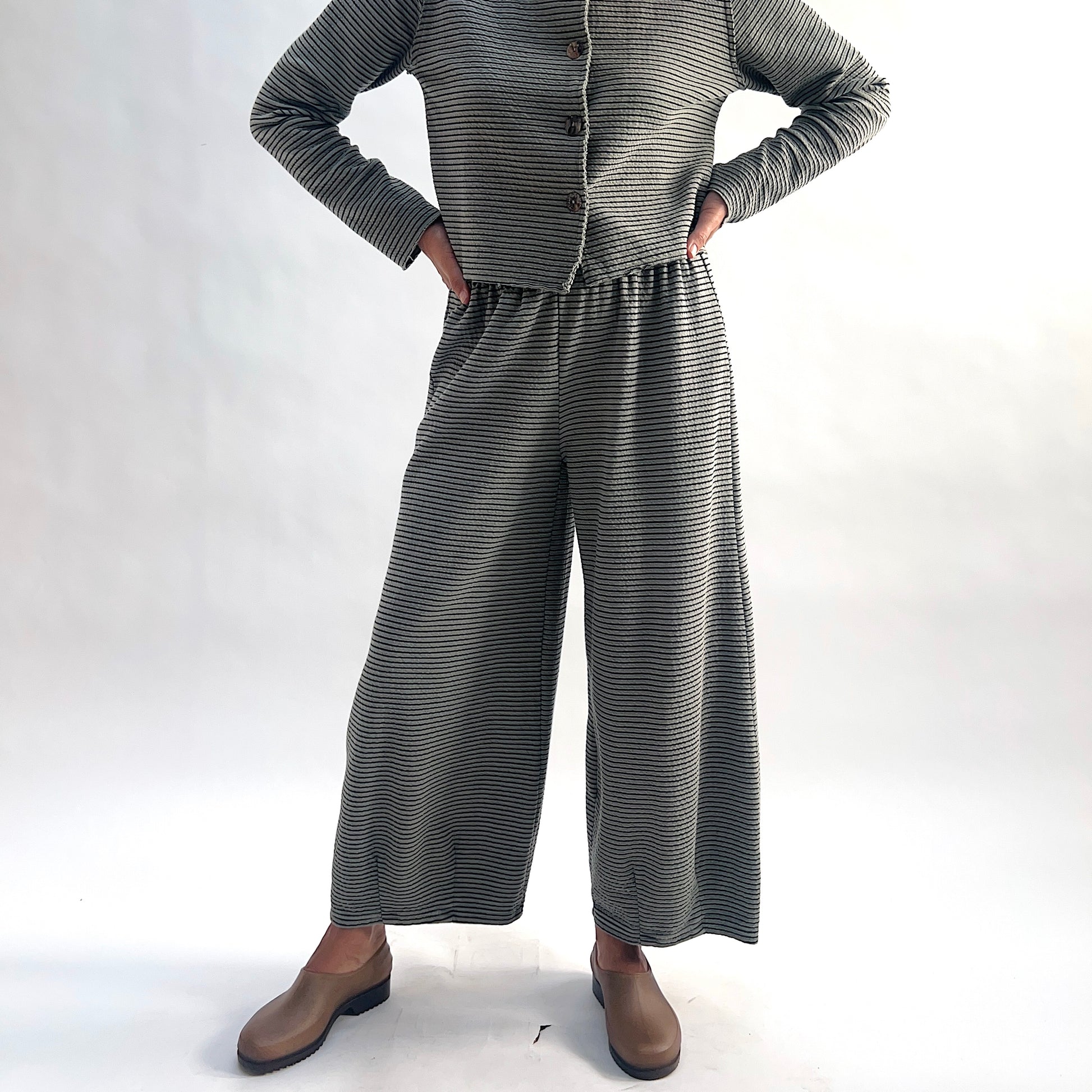 Full front view of model wearing squirrel and black fine horizontal stripe wide-leg cropped pants, hands on hips, showing the relaxed wide-leg silhouette and crinkle-texture stripe, styled with the matching striped asymmetrical cardigan and taupe slip-on shoes against a white background.
