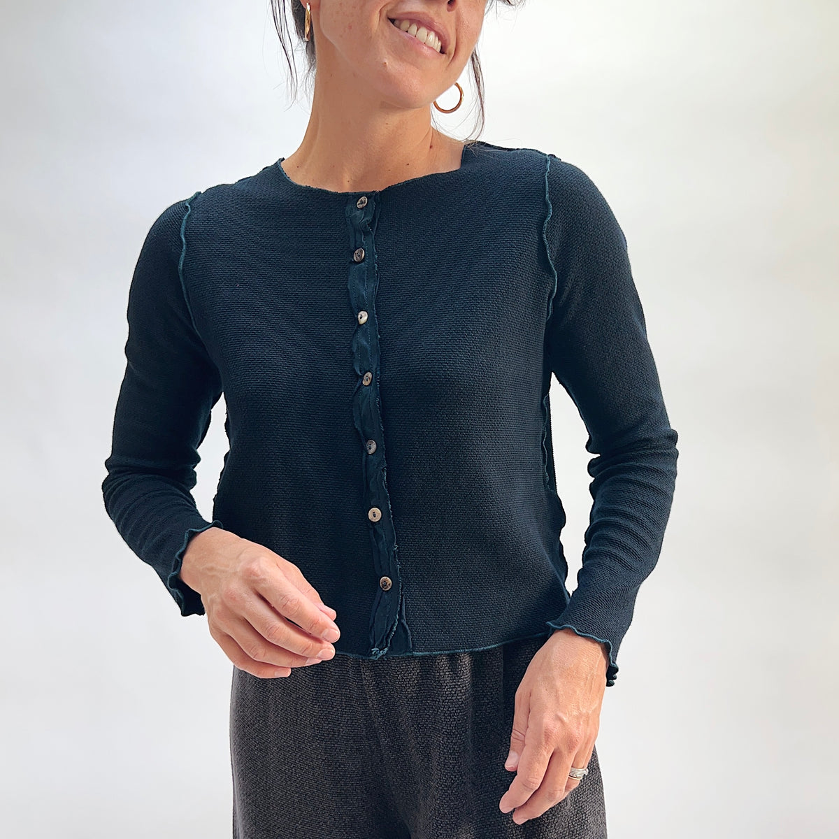 Model wearing the Cut Loose Marled Knit Cropped Cardigan in Etan, smiling close-up front view, deep teal marled knit cropped cardigan with scalloped lettuce-edge neckline, iridescent shell buttons, and long sleeves, styled with a dark textured skirt
