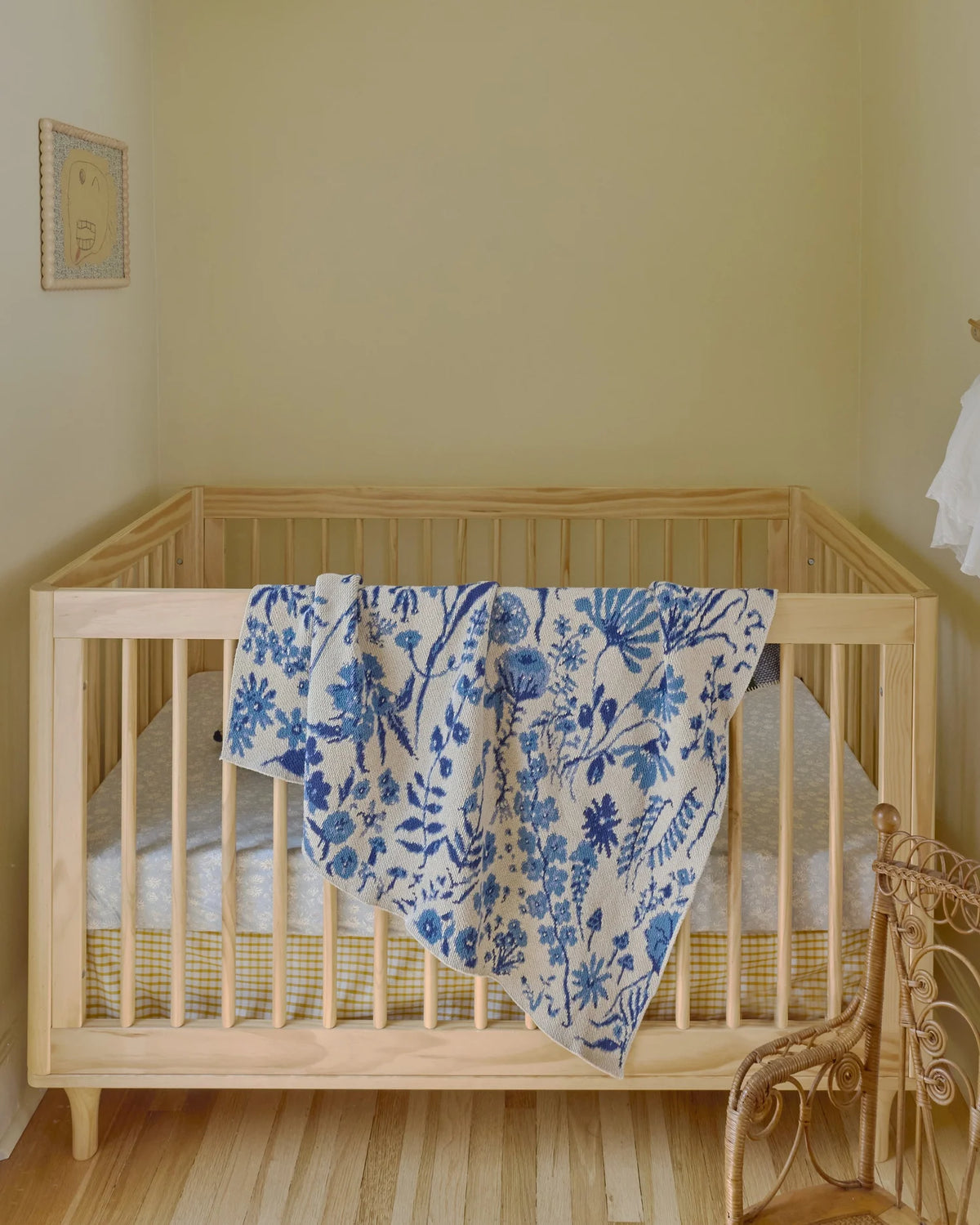 Flower Field Sky Blue blanket draped over a wooden crib rail, showing its blue floral pattern on cream.
