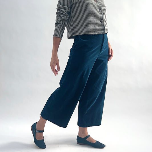 Three-quarter front view of model wearing deep teal mini cord straight crop trousers, leg extended mid-stride, showing the wide straight leg, flat front, and cropped length, styled with the matching white and black stripe asymmetrical cardigan and teal mary jane flats against a white background.
