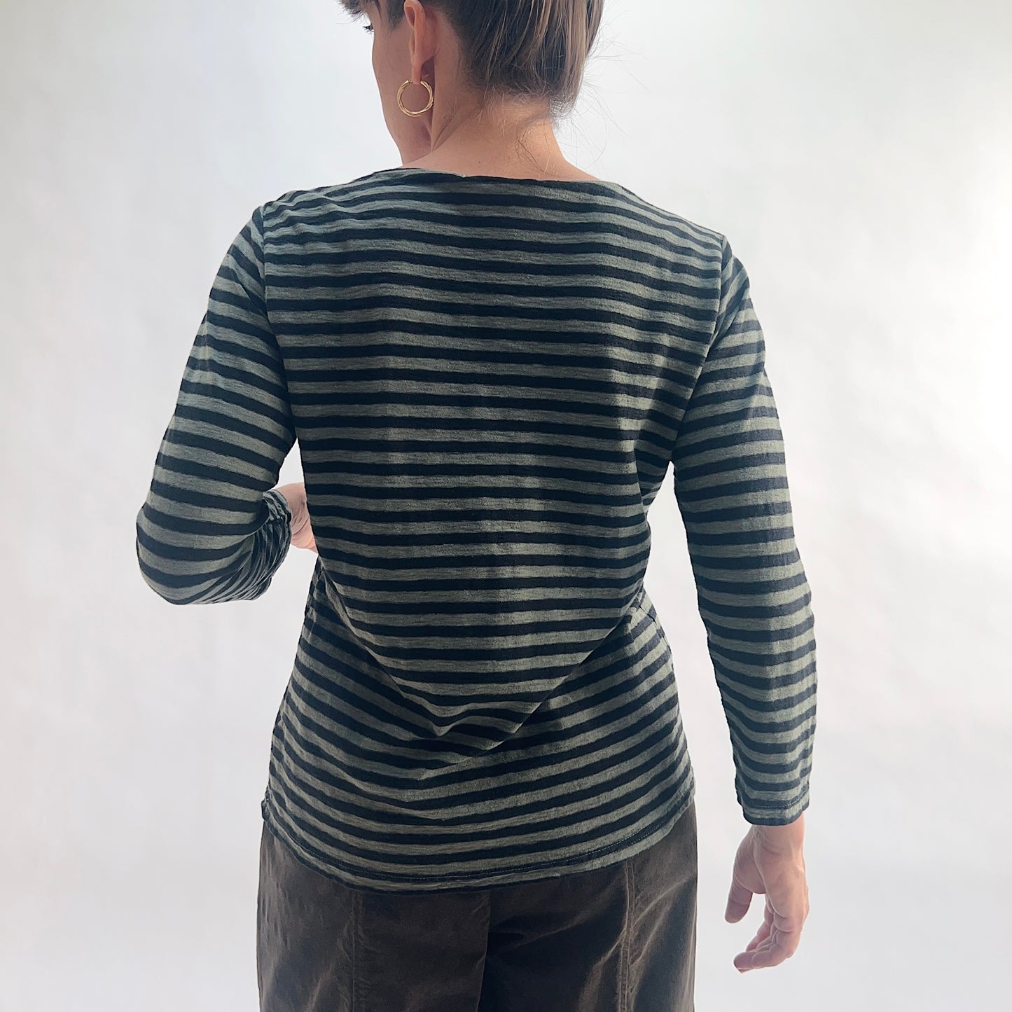 Back view of model wearing a kelp green and black horizontal stripe boatneck tee with 3/4 sleeves, hands behind back, showing the wide boatneck, linen-blend texture, and straight hem, styled with dark charcoal wide-leg pants against a white background.
