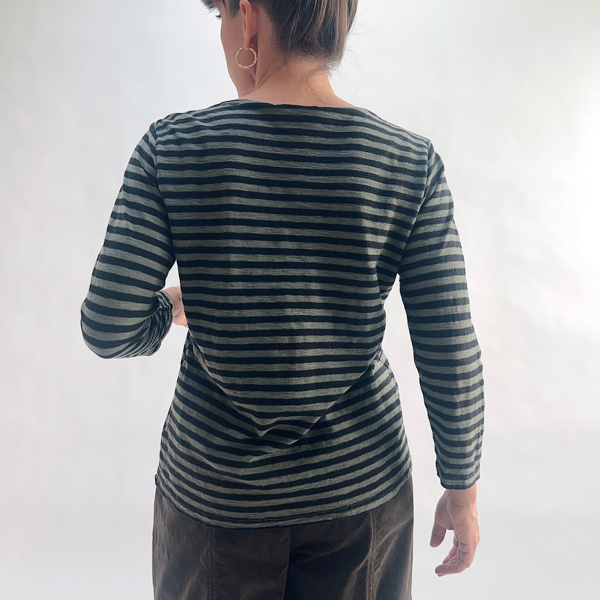 Back view of model wearing a kelp green and black horizontal stripe boatneck tee with 3/4 sleeves, hands behind back, showing the wide boatneck, linen-blend texture, and straight hem, styled with dark charcoal wide-leg pants against a white background.
