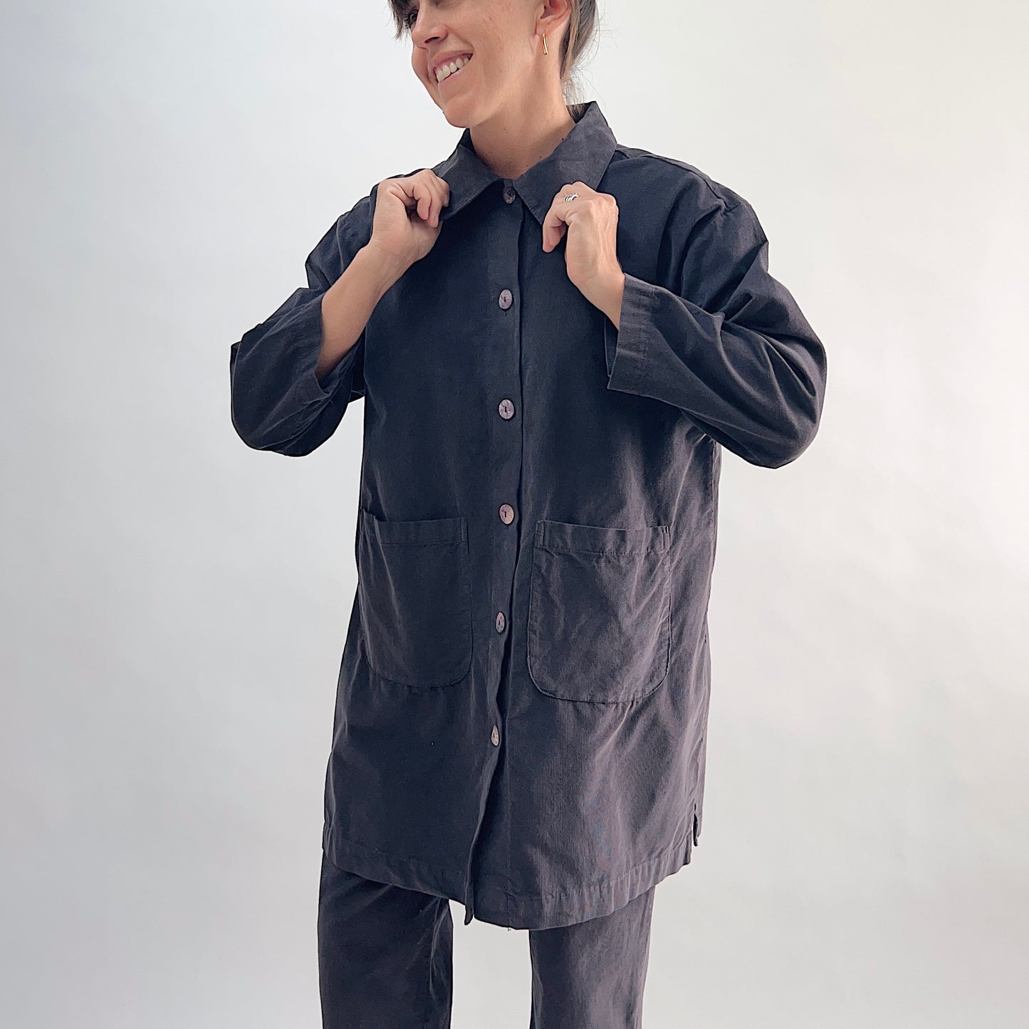 Model adjusting collar of the Bryn Walker Corduroy Car Coat in Fig, front view highlighting rounded point collar, mother of pearl button closure, two large patch pockets, 3/4 sleeves, side hem slits, and deep fig pinwheel corduroy, styled with matching corduroy pants
