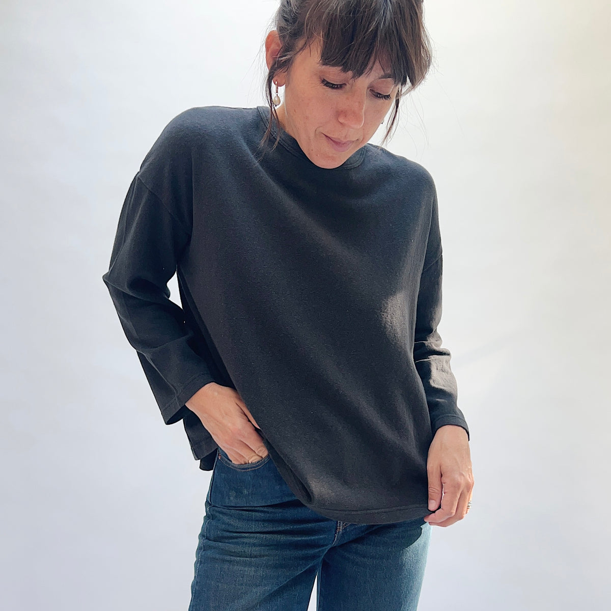 Model looking down while wearing the black cotton knit pullover with blue jeans and holding the front hem.
