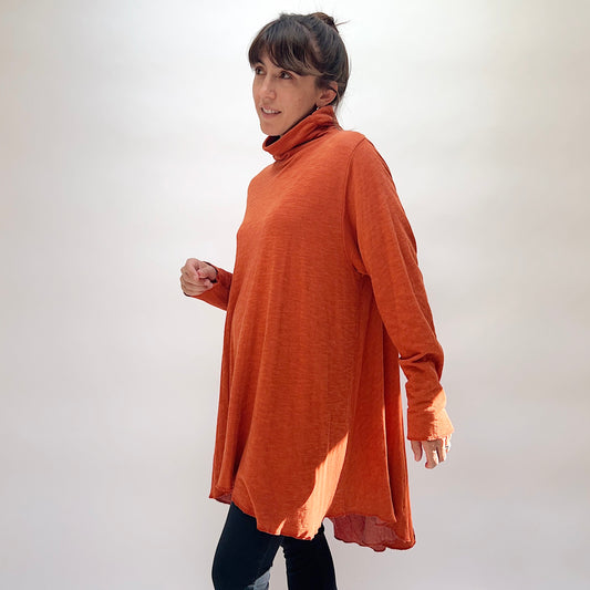 North Star Base Double Cotton Hi Low Turtleneck Top in Raw Sienna, side view on smiling model in motion, raw sienna orange double-layered cotton with draped turtleneck collar, long sleeves with raw cuffs, and sweeping hi-low hem with raw edge, paired with dark jeans
