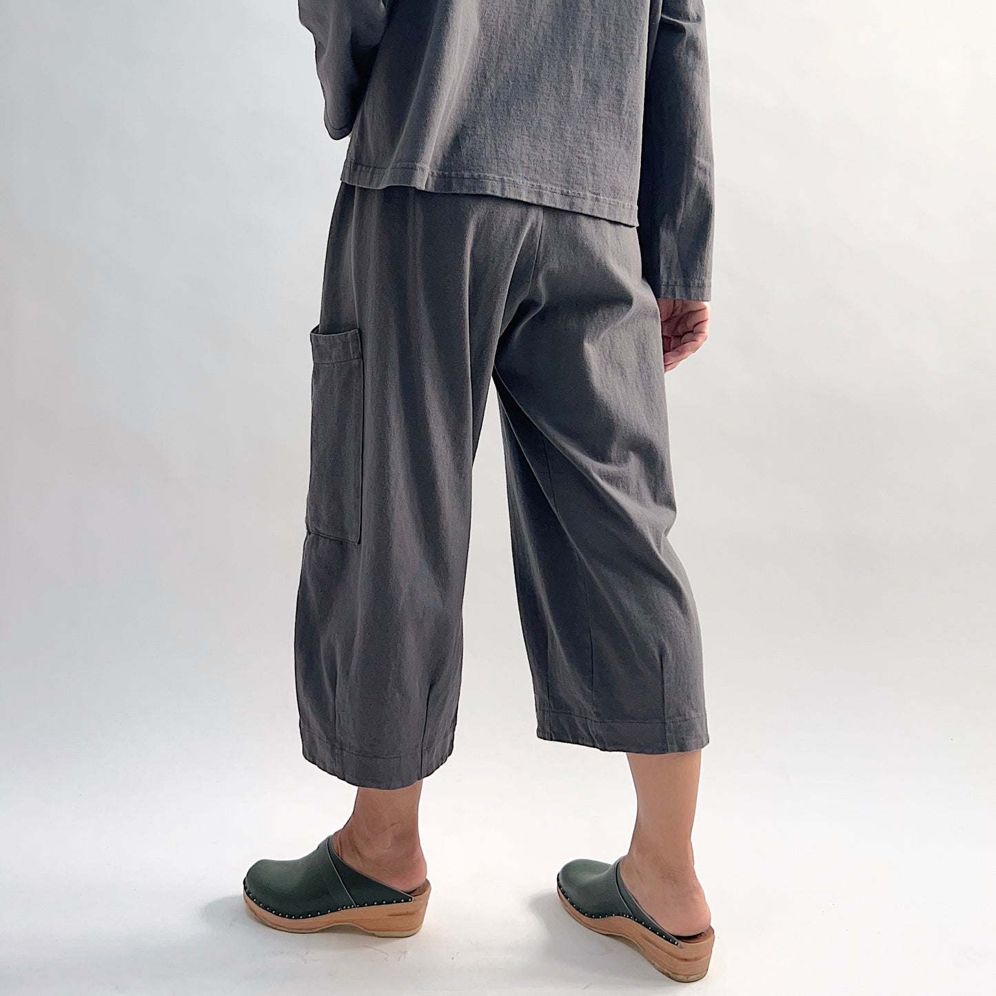 Back view of model wearing the Bryn Walker Cotton Pasha Pant in Moonlight, showing elastic gathered waistband, wide-leg cropped silhouette, thigh patch pocket, folded cuff hem, and soft blue-grey cotton, styled with matching top and green wedge clogs
