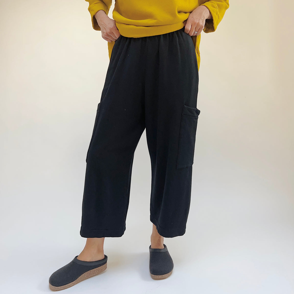 Front lower-body view of a woman wearing the Bryn Walker Pasha Fleece Pant in Black, showing the elastic waistband, two large thigh patch pockets, wide straight leg silhouette, and folded ankle hem in black organic cotton and bamboo fleece, styled with a mustard yellow sweatshirt and dark slide clogs against a white background
