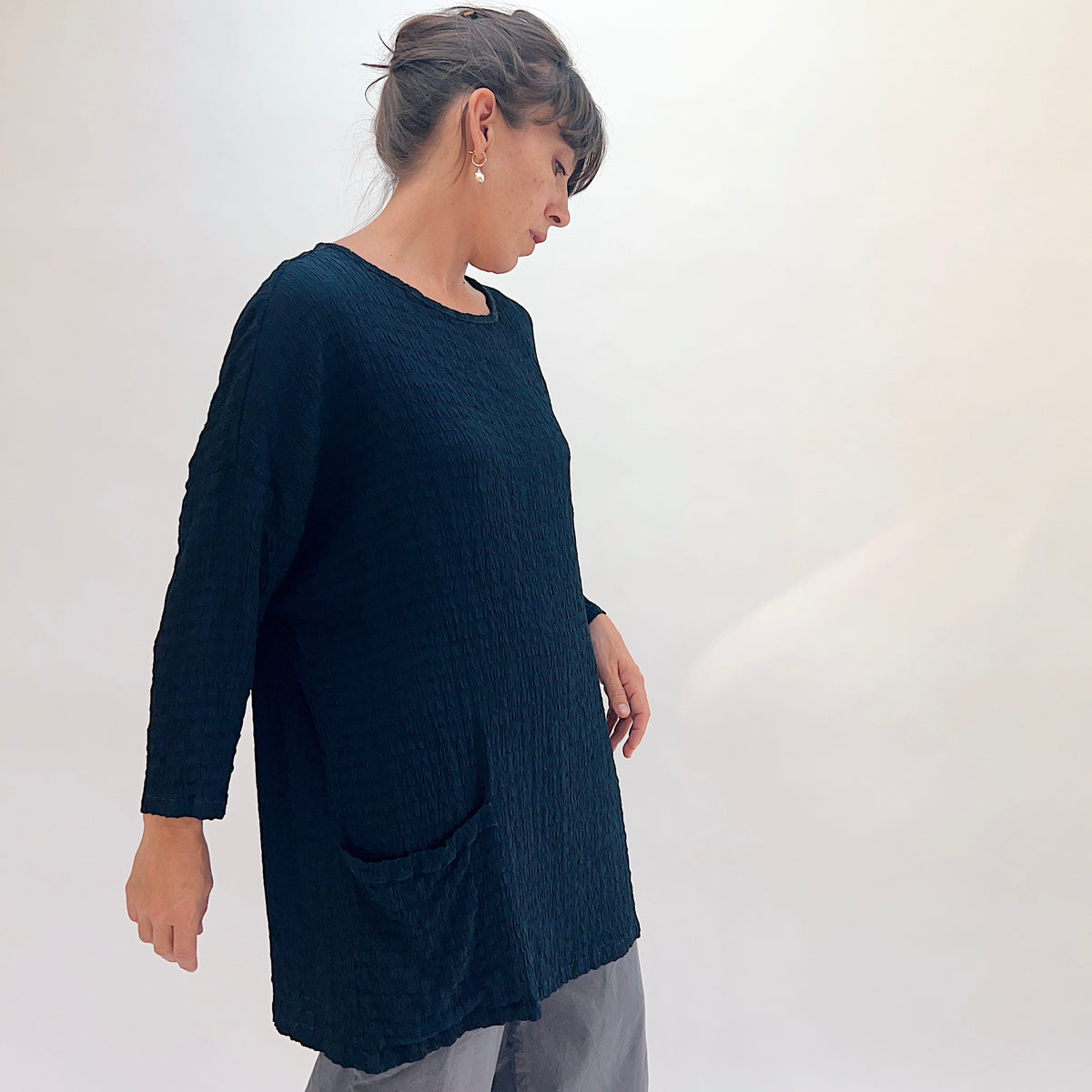 Front view of teal crinkle-texture one-size pocket tunic showing scoop neckline, relaxed fit, 3/4-length sleeves, and two front patch pockets, paired with grey wide-leg pants
