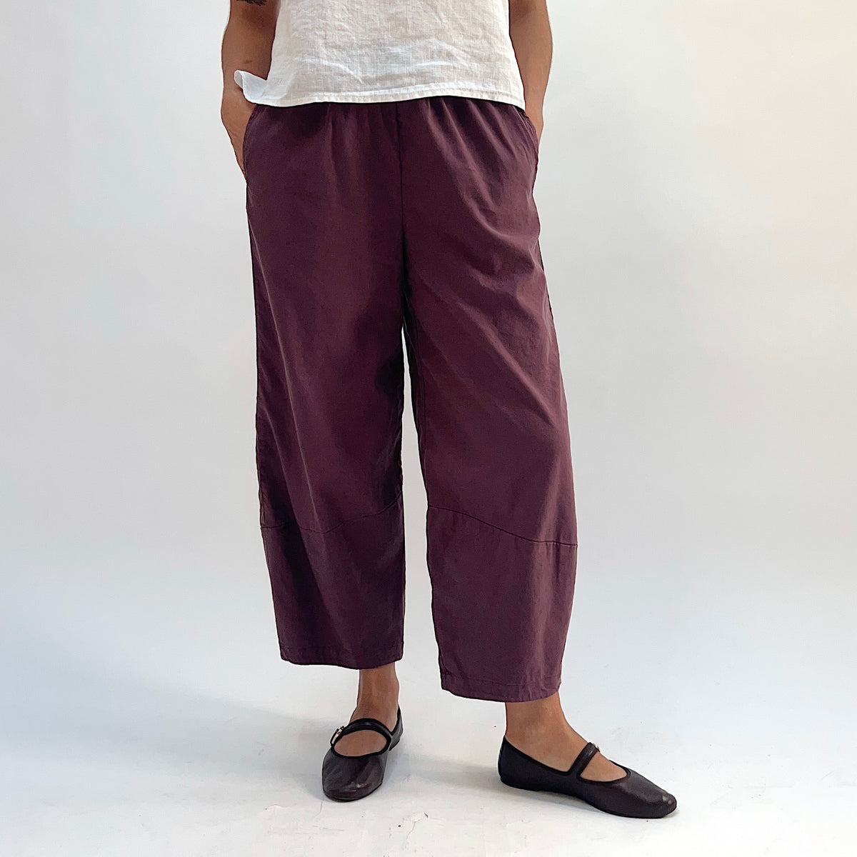 Full front view of russet linen-tencel lantern pants with wide barrel-leg silhouette, elastic waistband, side pockets, and curved hem, styled with a white linen top and dark Mary Jane flats
