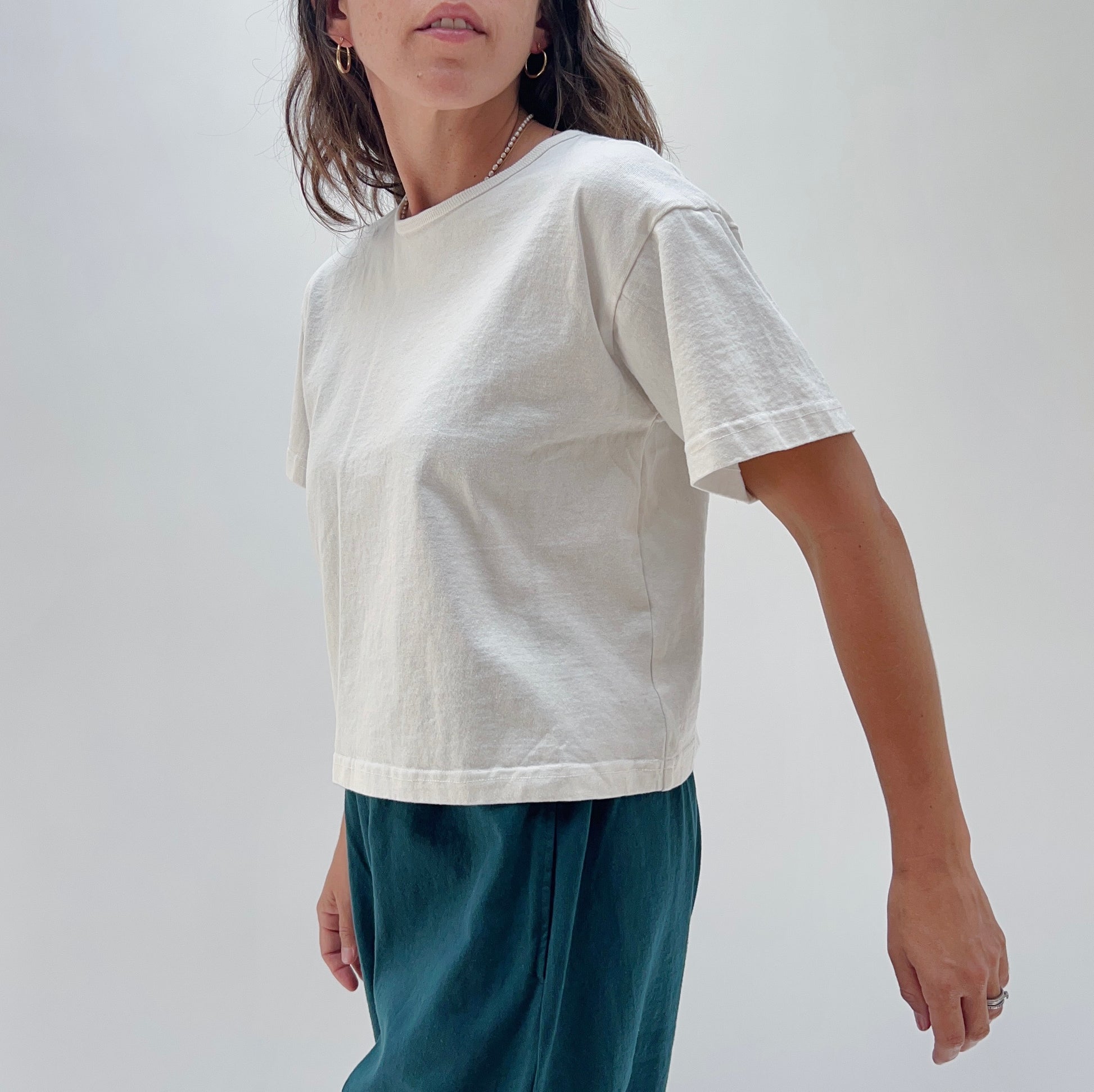 A person wearing a white, short-sleeved, cropped t-shirt and dark pants. The image focus is on the t-shirt.