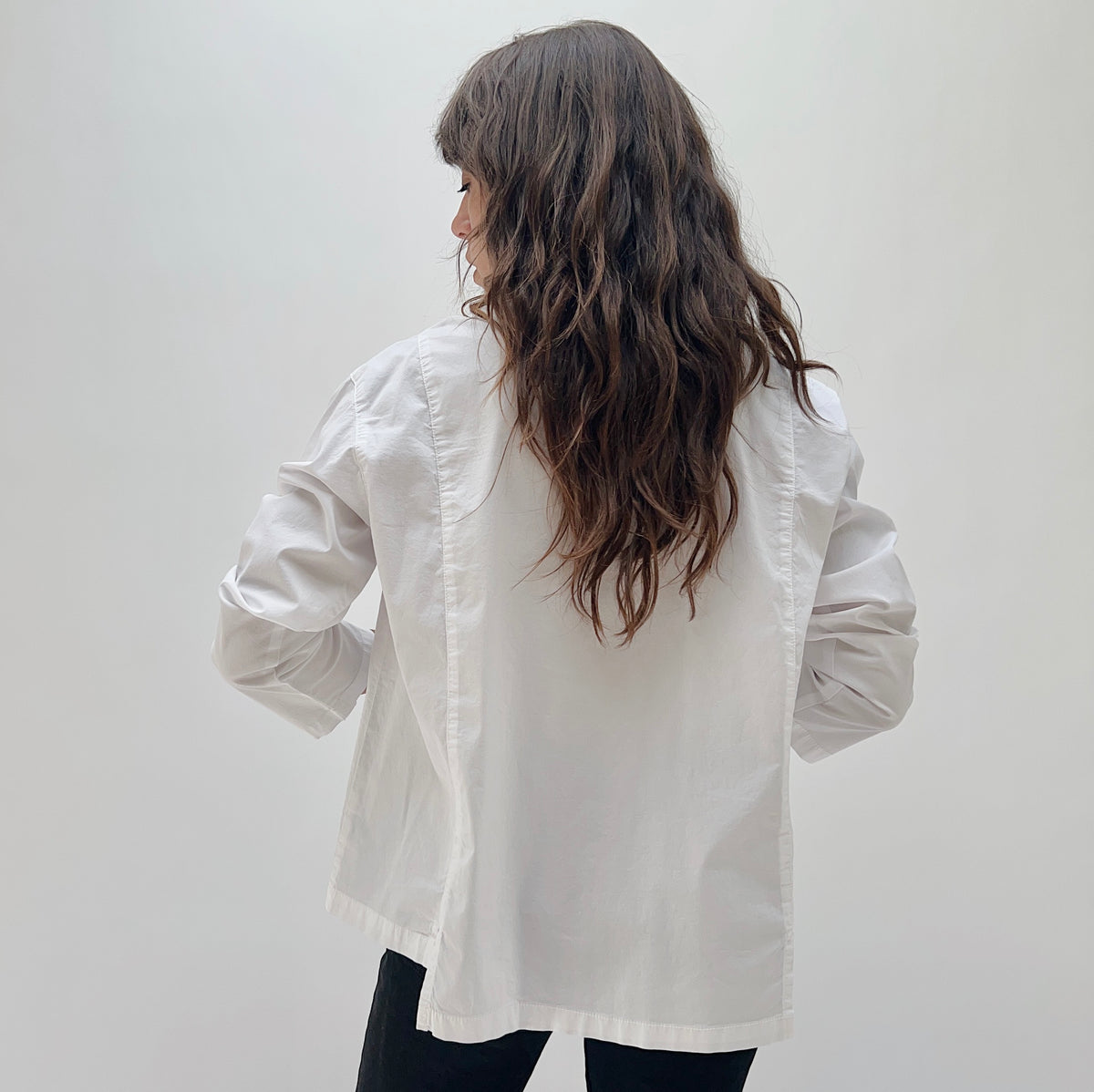 Model wearing the Eleven Stitch Tuxedo Shirt in Chalk, back view, showing clean seaming and relaxed boxy fit of white cotton tuxedo shirt, styled with black pants

