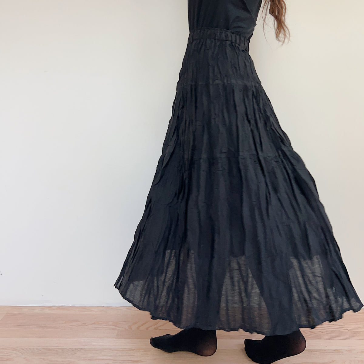 Side view of the black pleated skirt on a model, showing the elastic waistband and flowing hem.
