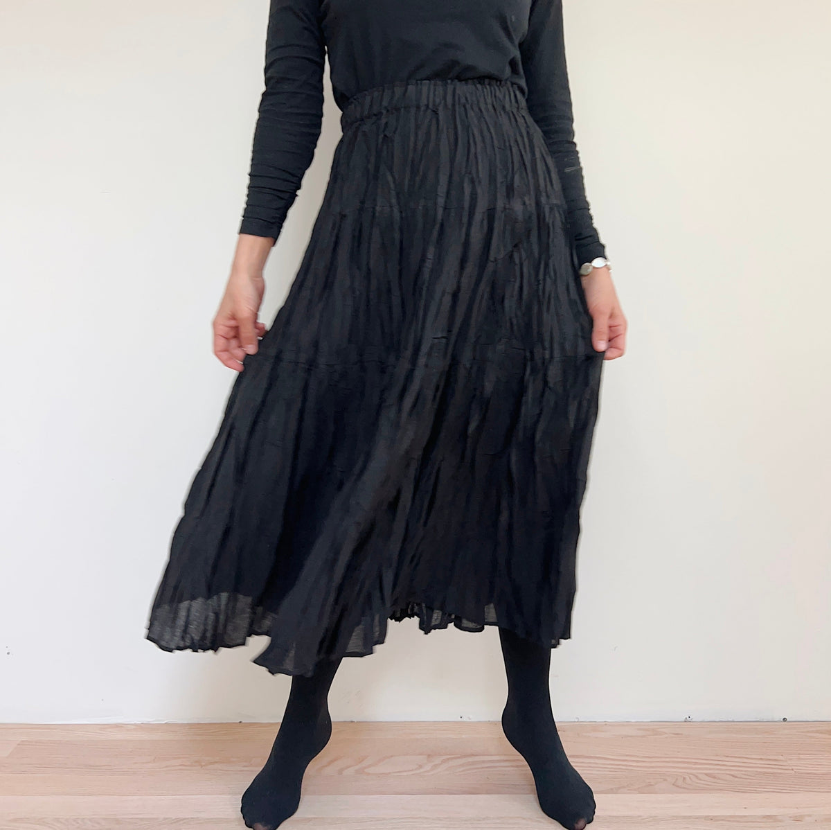 Front view of the black pleated skirt on a model, holding the sides to show its drape.
