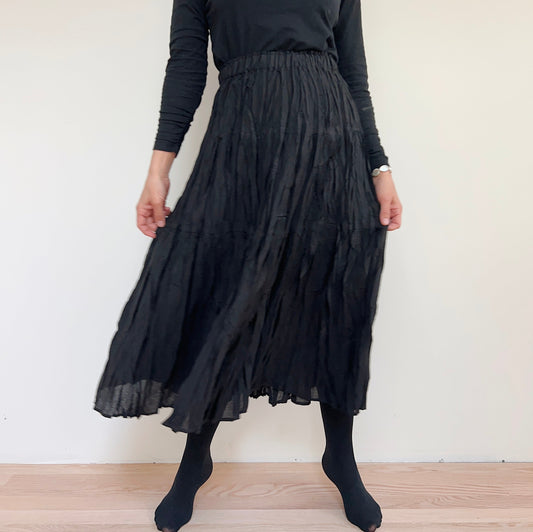Front view of the black pleated skirt on a model, holding the sides to show its drape.
