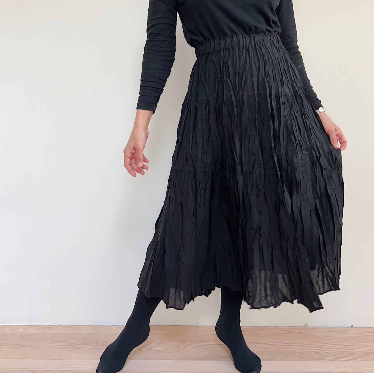 Front view of the black pleated skirt on a model with one leg extended, showing its length and textured pleats.
