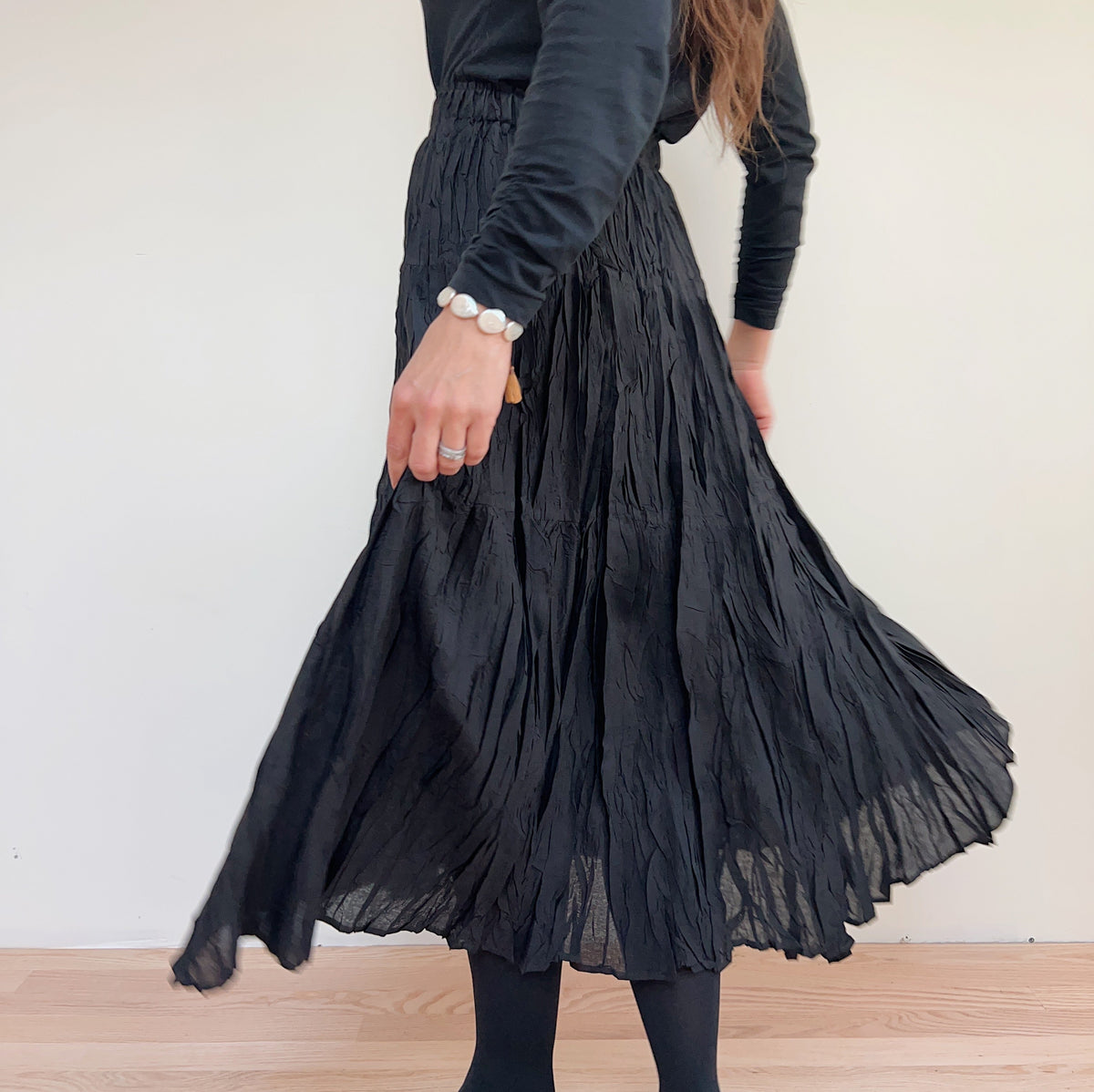Three-quarter view of the black pleated skirt on a model turning, with the hem flaring outward.
