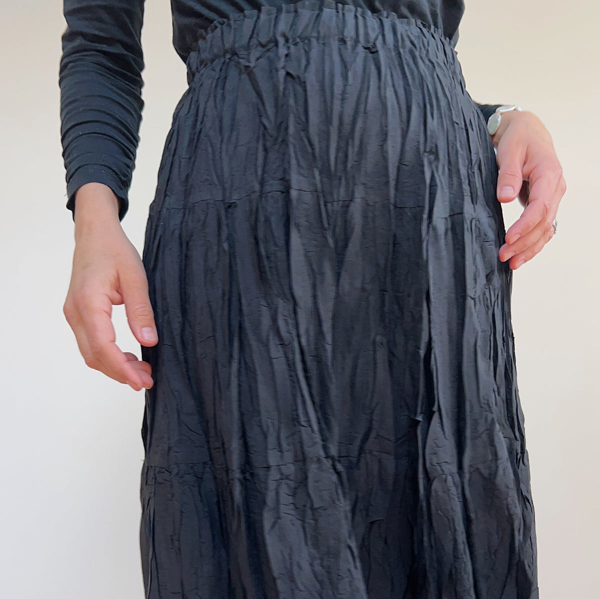 Close-up of the black pleated skirt on a model, showing the gathered elastic waistband and crinkled fabric texture.
