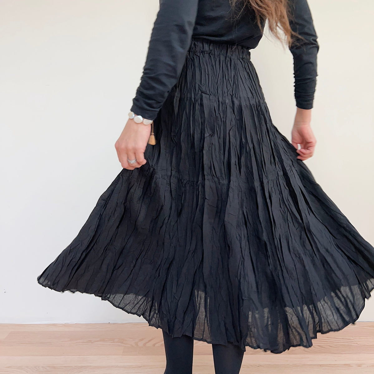 Rear three-quarter view of the black pleated skirt on a model turning, showing the wide, flared hem.

