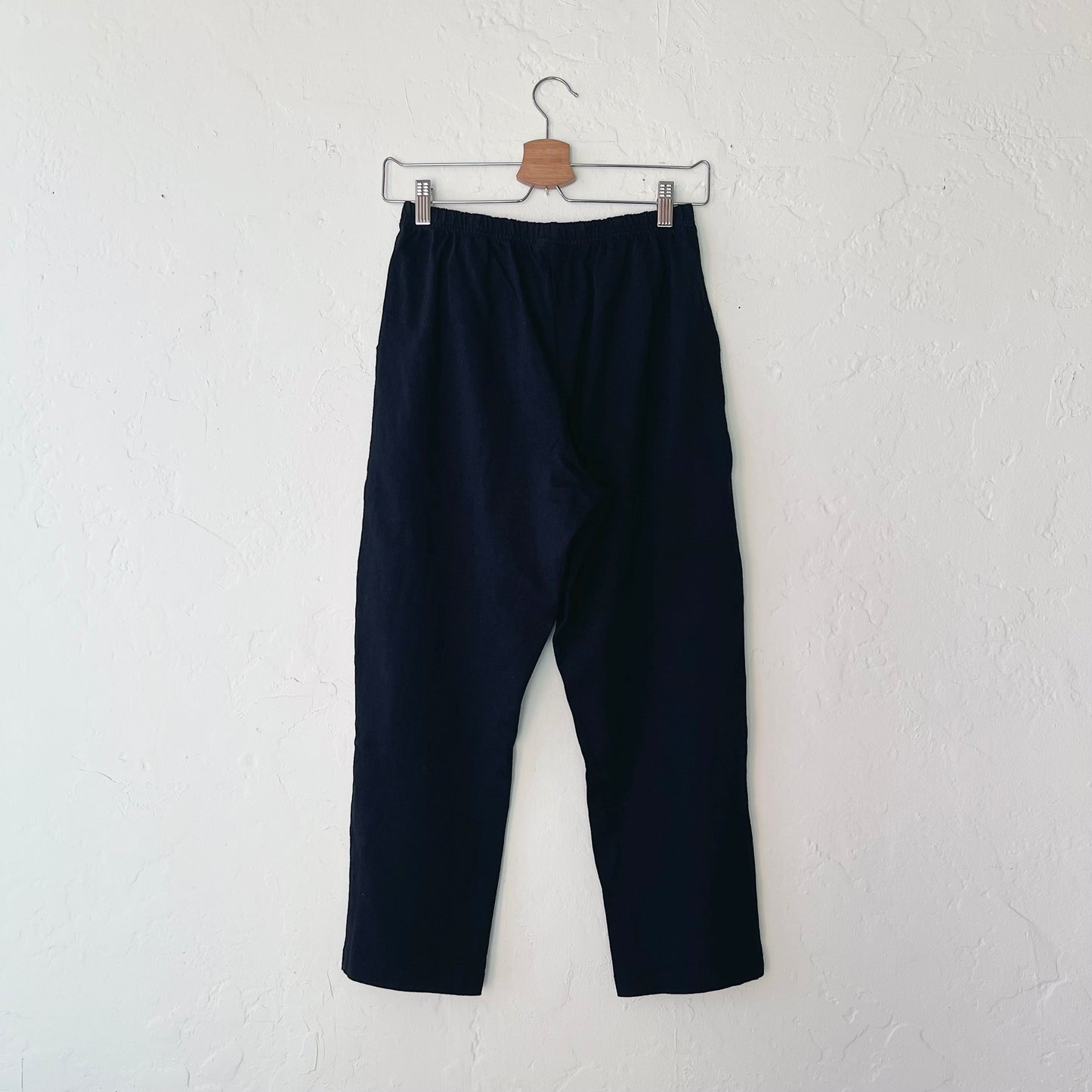 A pair of black cotton pants hanging on a hanger against a white wall.