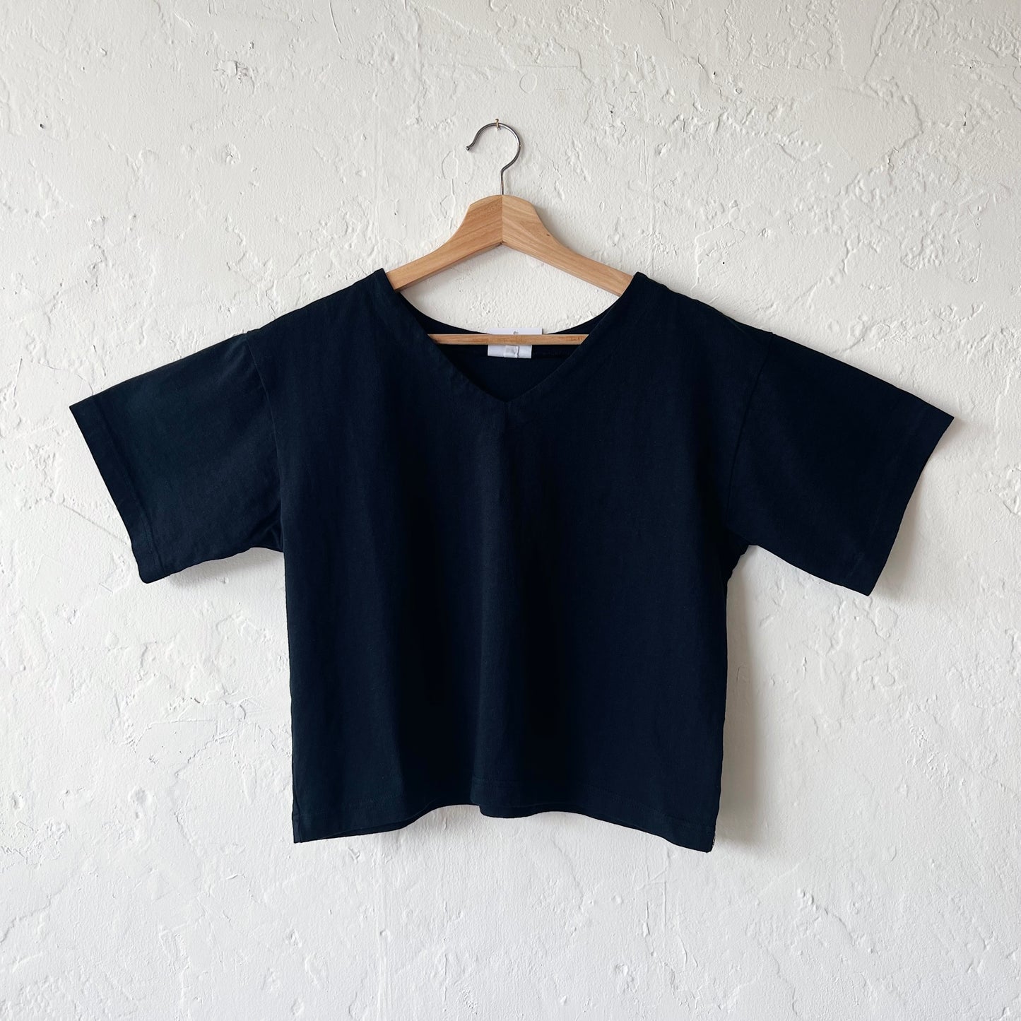 A black, cropped sleeve top with a slight v-neck hanging on a hanger against a white textured wall.