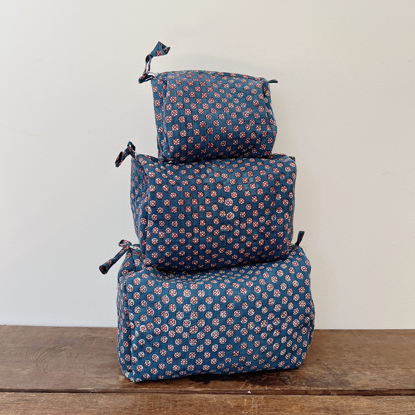 Three sizes of the Toiletry Bag in Alma Blockprint stacked on a wooden surface — small, medium, and large — in a navy blue quilted cotton with a pink and white floral block-print pattern