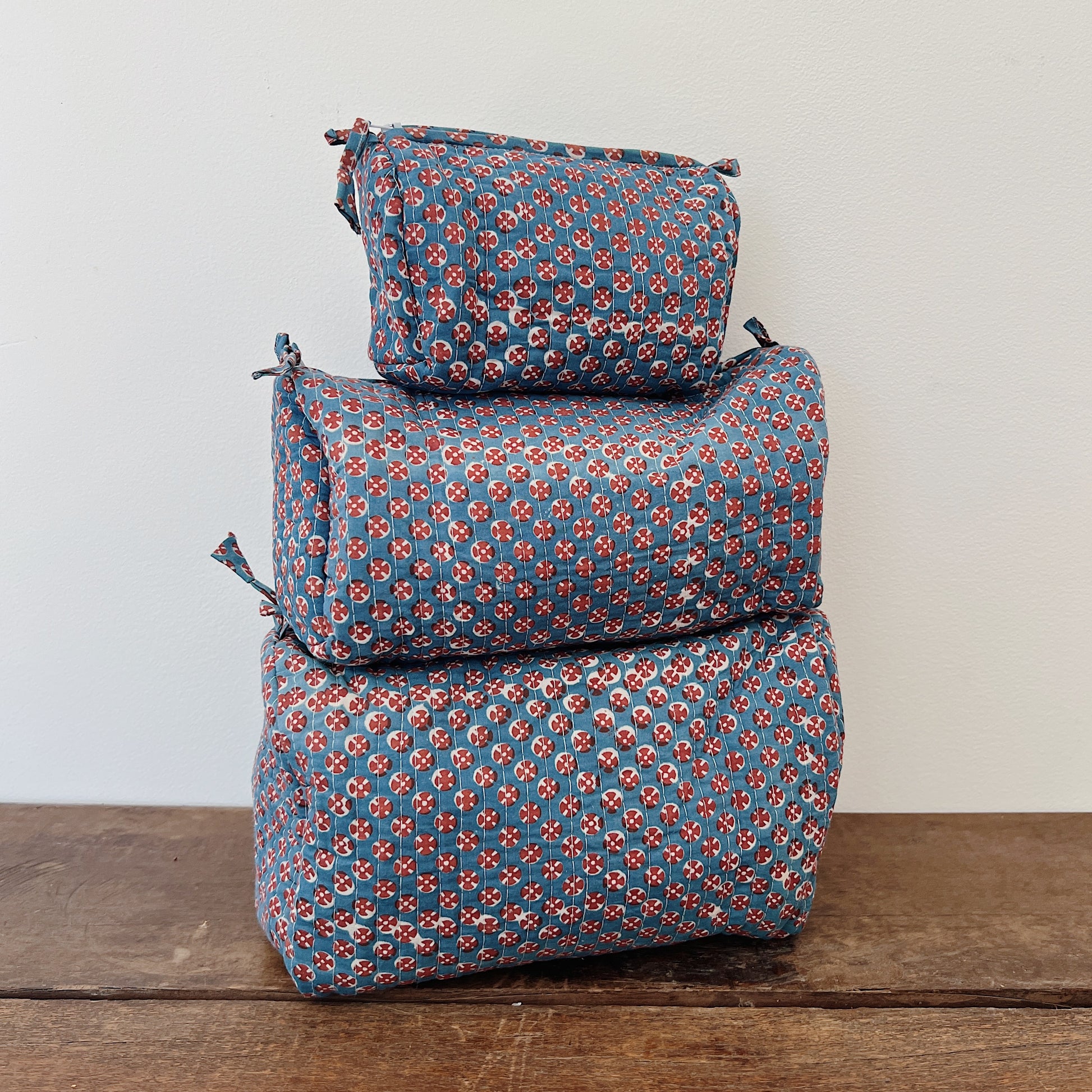 Three sizes of a quilted block-print toiletry bag stacked on a wooden surface — small, medium, and large — in a slate blue cotton with a red and cream circular block-print pattern