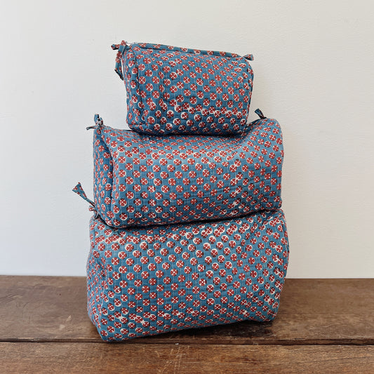 Three sizes of a quilted block-print toiletry bag stacked on a wooden surface — small, medium, and large — in a slate blue cotton with a red and cream circular block-print pattern