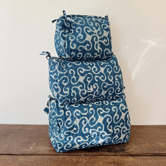 hree sizes of the Toiletry Bag in Demeter Blockprint stacked on a wooden surface — small, medium, and large — in a deep indigo quilted cotton with a white scrollwork block-print pattern