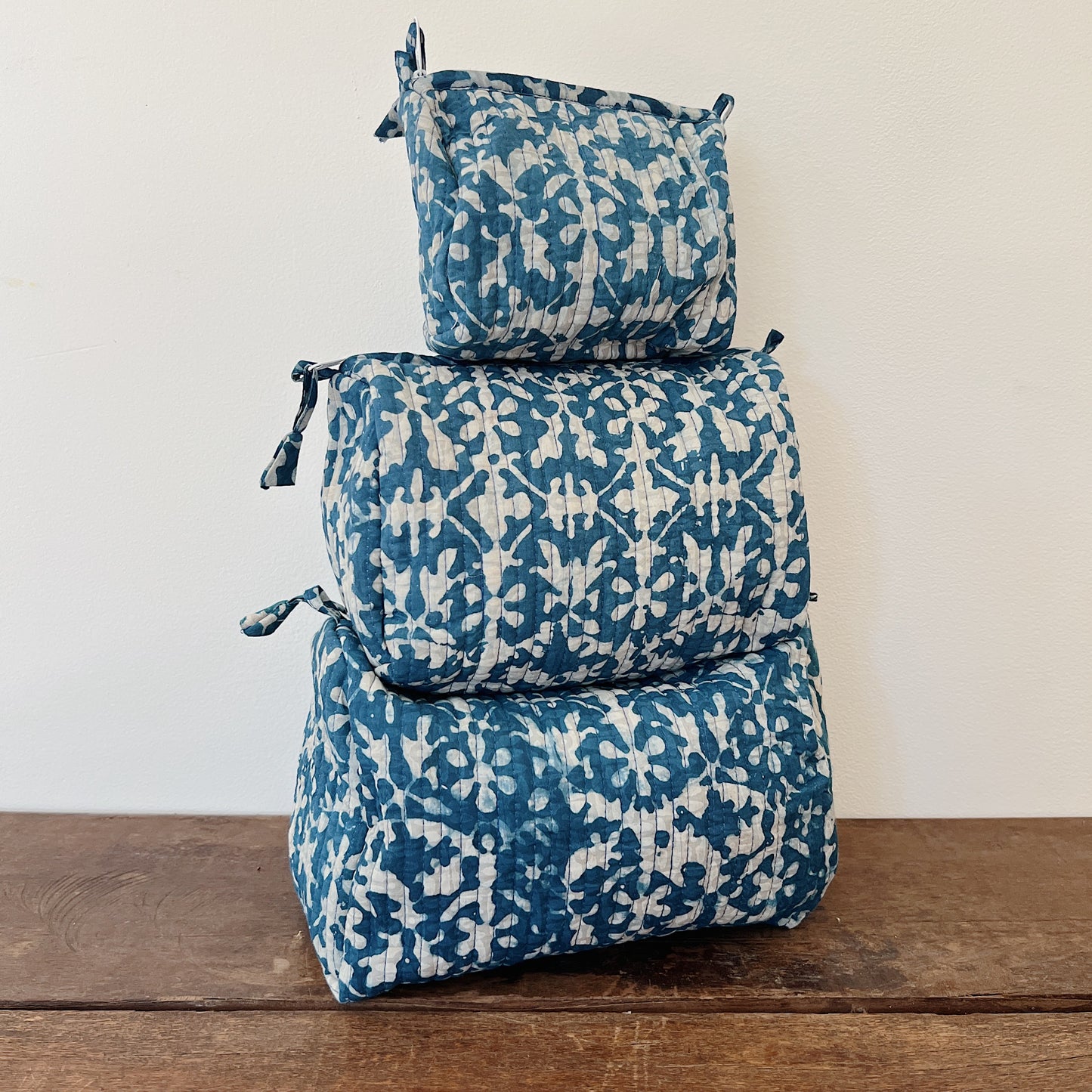 Three sizes of the Toiletry Bag in Isla Blockprint stacked on a wooden surface — small, medium, and large — in a cream quilted cotton with a teal floral tile block-print pattern