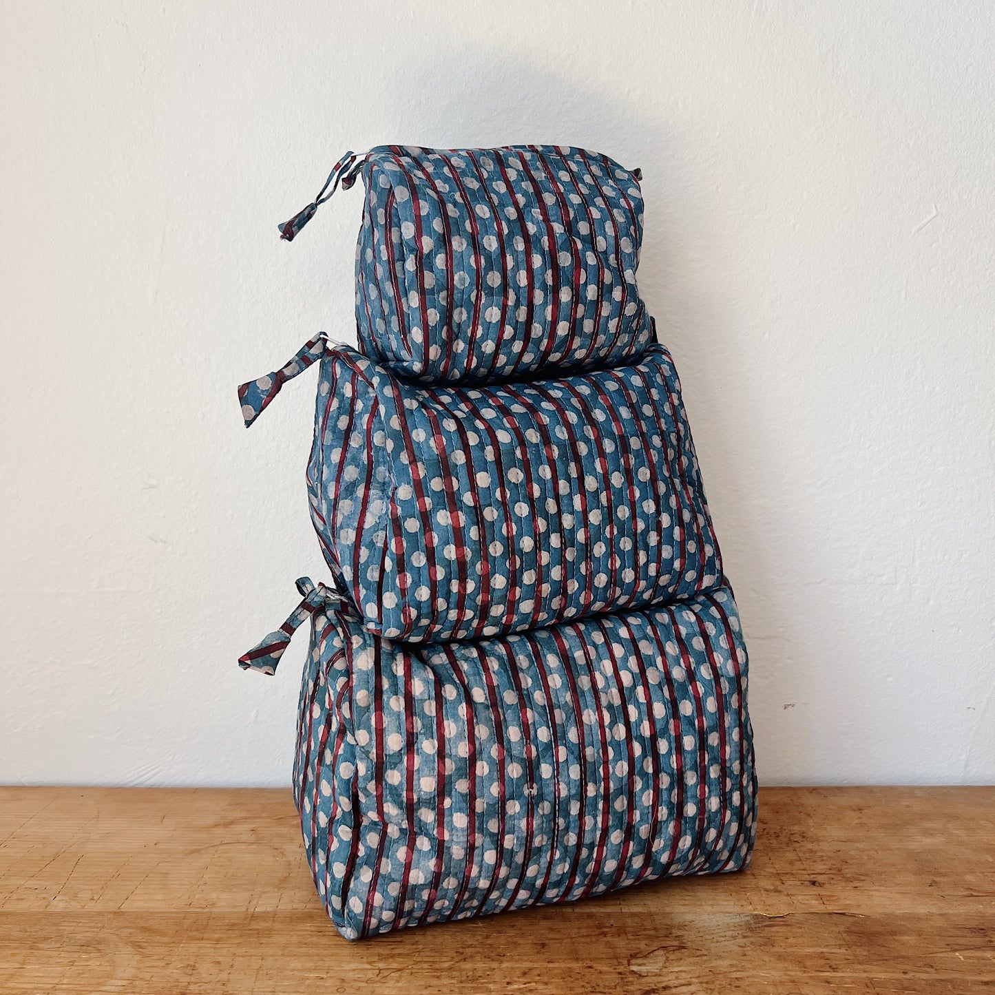Three sizes of the Toiletry Bag in Persephone Blockprint stacked on a wooden surface — small, medium, and large — in a slate blue quilted cotton with burgundy stripes and white polka dot block-print pattern