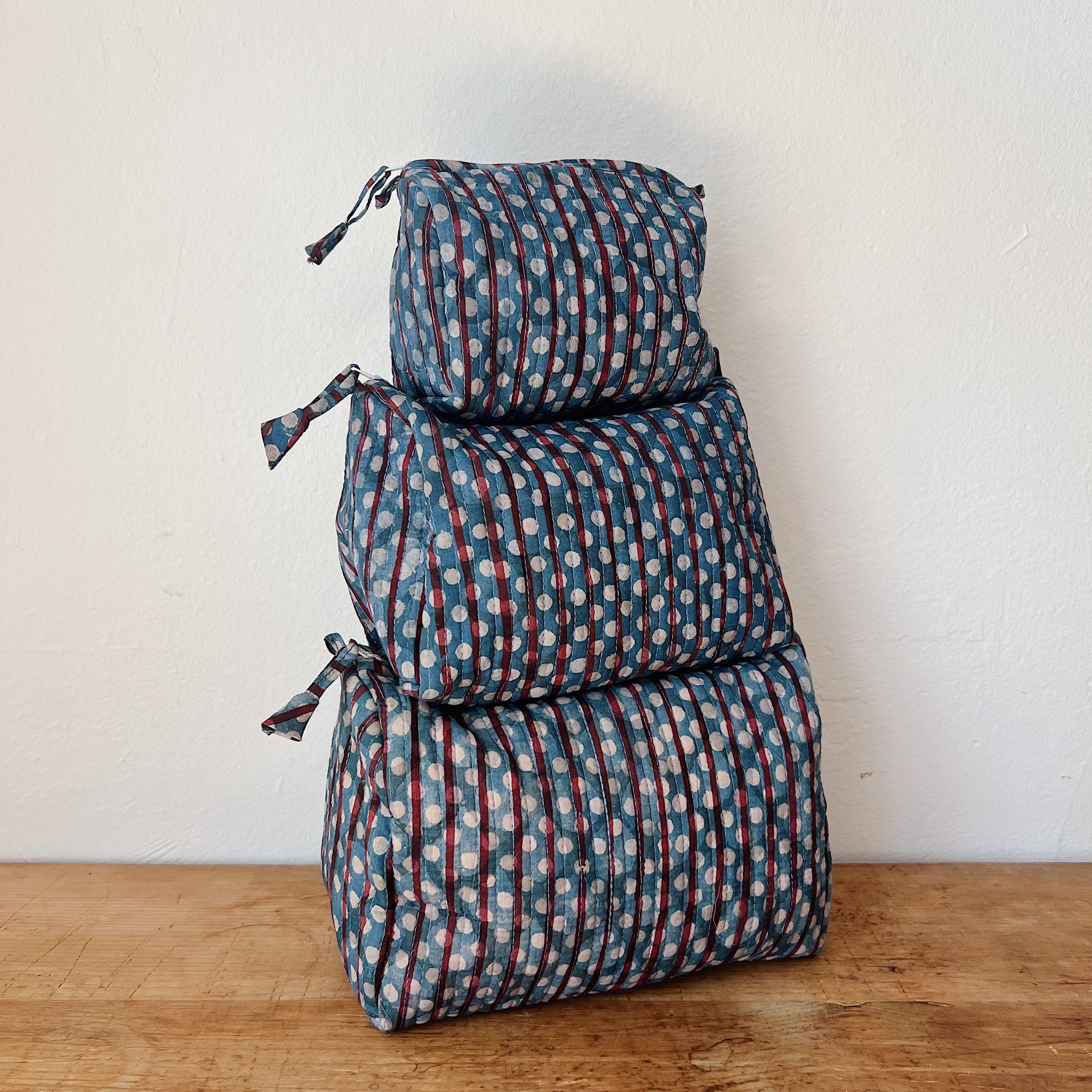 Three sizes of the Toiletry Bag in Persephone Blockprint stacked on a wooden surface — small, medium, and large — in a slate blue quilted cotton with burgundy stripes and white polka dot block-print pattern
