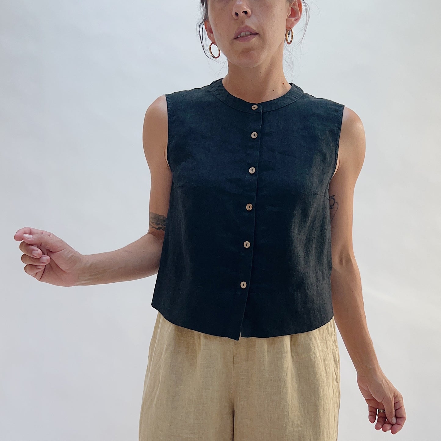 Three-quarter front view of the Button Front Tank in Black by Yuvita, showing the cropped linen silhouette, button-front detail, and collarless neckline, styled with tan linen trousers