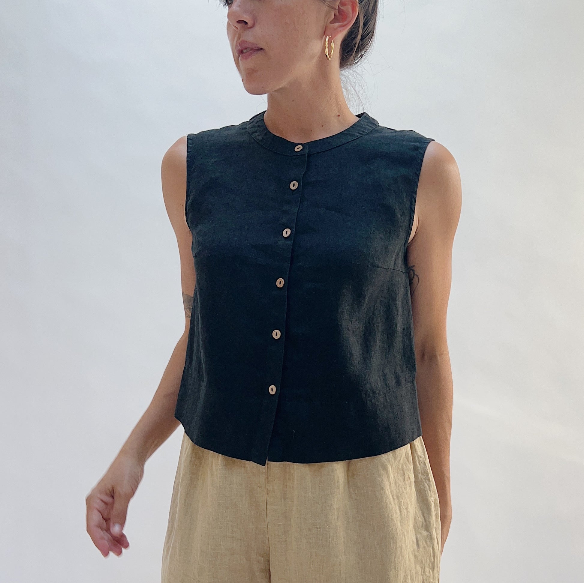 Front close-up of the Button Front Tank in Black by Yuvita, showing the full button-front placket, collarless neckline, and black linen fabric, styled with tan linen trousers
