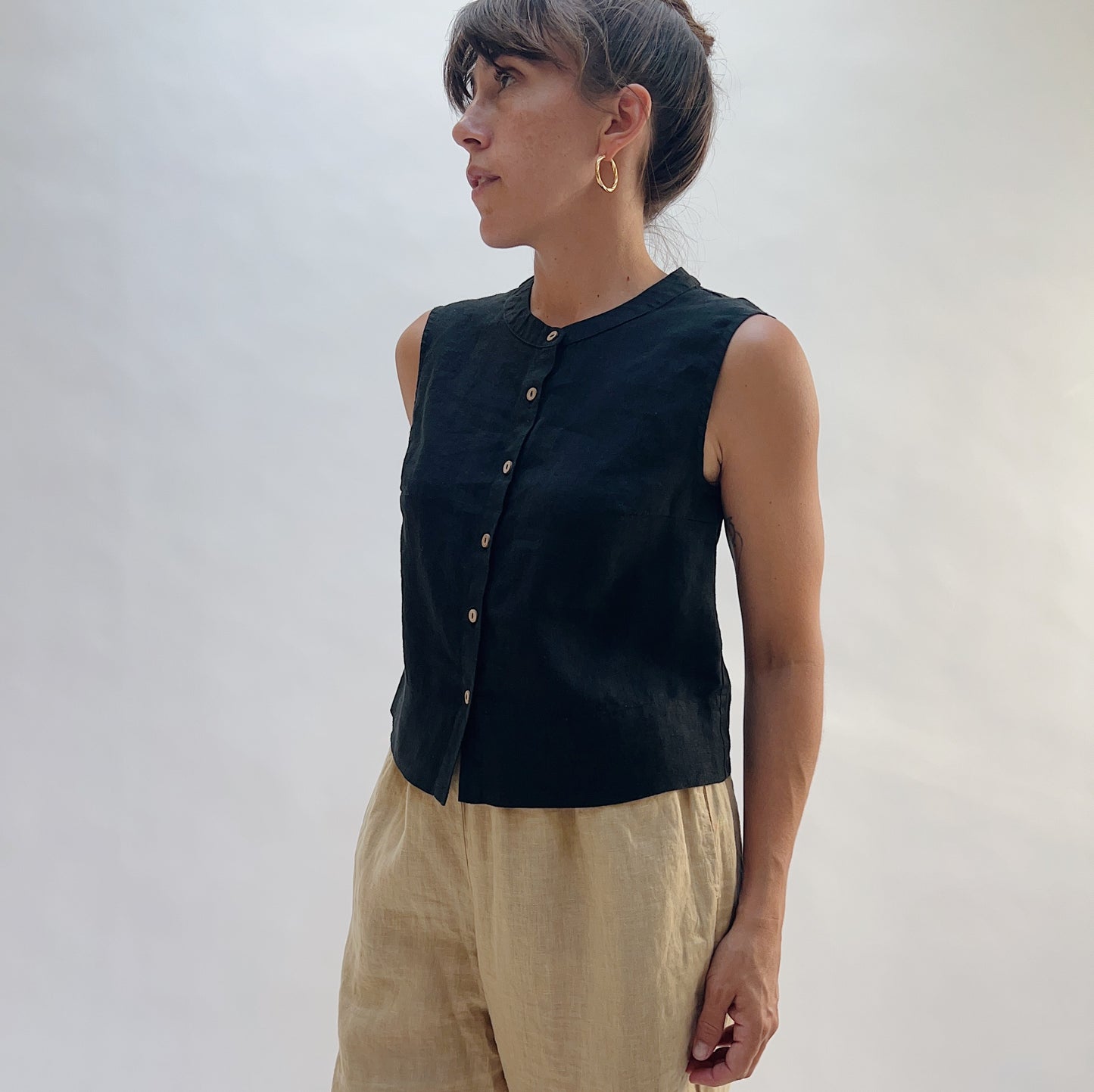 Front view of the Button Front Tank in Black by Yuvita — a cropped 100% linen collarless button-front tank in black, styled with tan linen trousers on a white background