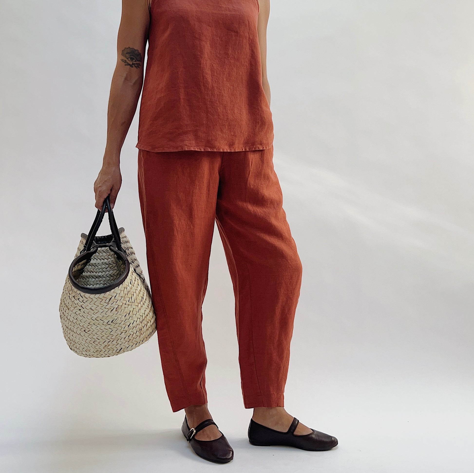 Three-quarter view of the Seamed Taper Pant in Terracotta by Yuvita, showing the tapered linen silhouette and seamed front leg, styled with a matching terracotta linen tank, woven bag, and dark Mary Jane flats