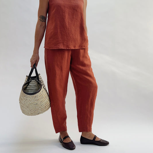 Three-quarter view of the Seamed Taper Pant in Terracotta by Yuvita, showing the tapered linen silhouette and seamed front leg, styled with a matching terracotta linen tank, woven bag, and dark Mary Jane flats