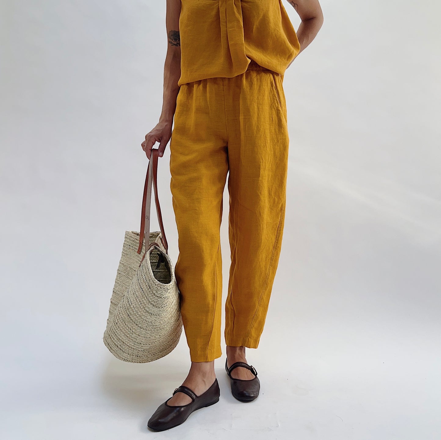 Three-quarter view of the Seamed Taper Pant in Tumeric by Yuvita, showing the tapered linen silhouette and seamed front leg in golden yellow, styled with a matching linen top, woven tote bag, and dark Mary Jane flats