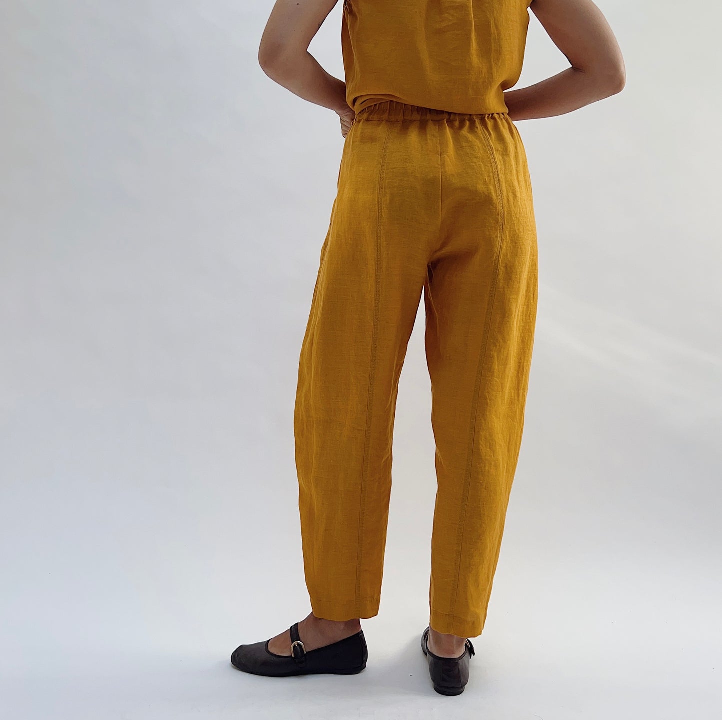 Back view of the Seamed Taper Pant in Tumeric by Yuvita, showing the elastic waistband, seamed back leg, and tapered ankle in golden yellow linen, styled with a matching linen top and dark Mary Jane flats
