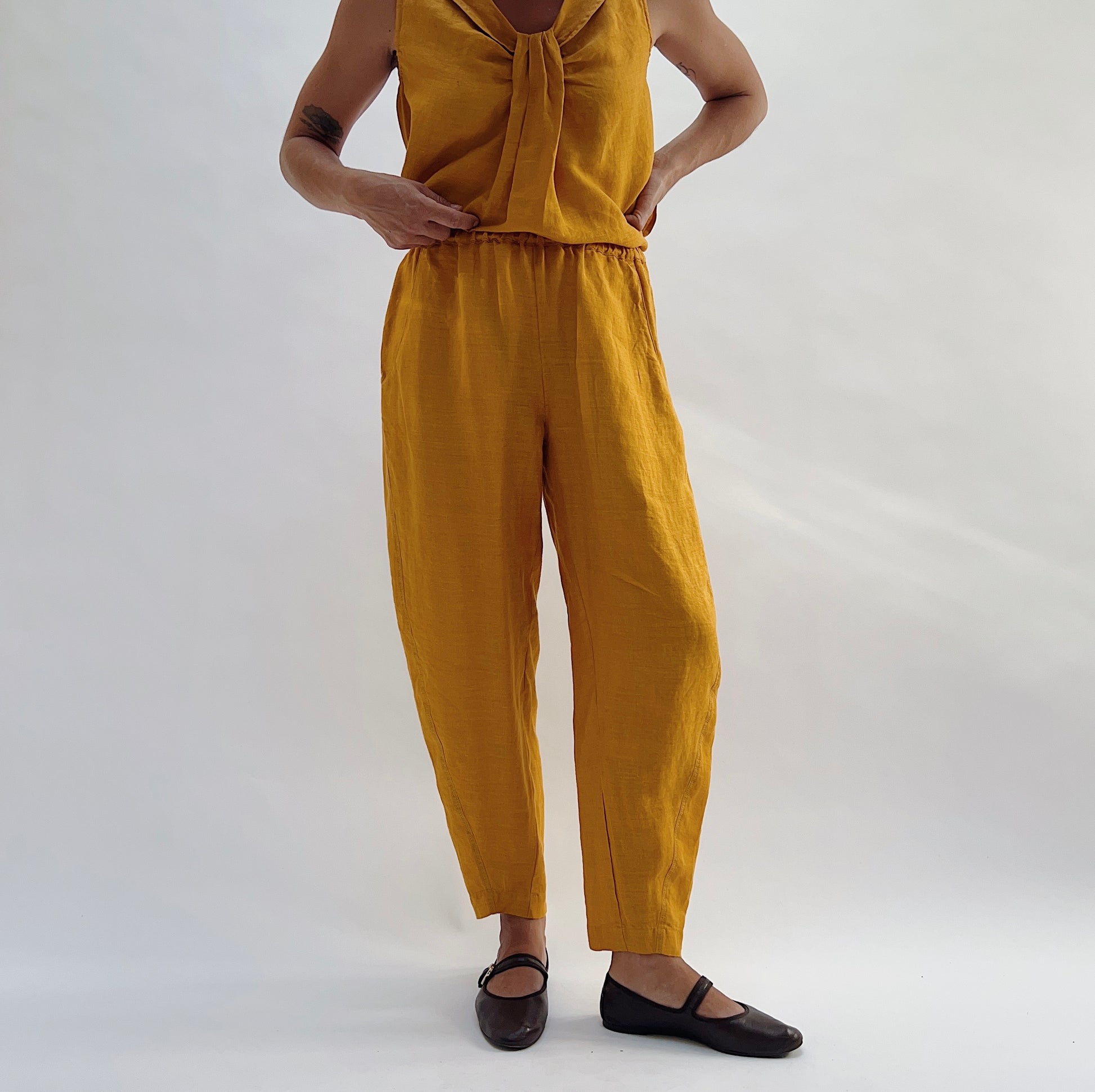 Front view of the Seamed Taper Pant in Tumeric by Yuvita, showing the elastic waistband, side pockets, and tapered ankle in golden yellow linen, styled with a matching tie-front linen top and dark Mary Jane flats